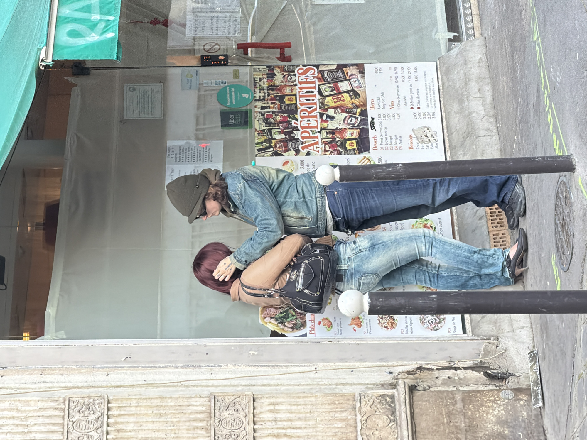 A street scene in Paris, France, shows two individuals standing in front of a restaurant or cafe. The person on the left wears a green hat, glasses, and a denim jacket, leaning in toward the person on the right, who has reddish-brown hair, a tan jacket, and a black purse. They are both standing behind a black bollard. Behind them, there is a glass window displaying menus, an Uber Eats sticker, and advertisements for aperitifs and other offerings. The street is visible on the right side of the frame, with a manhole cover and a yellow line marking. The lower portion of the frame shows a building facade with decorative carvings.