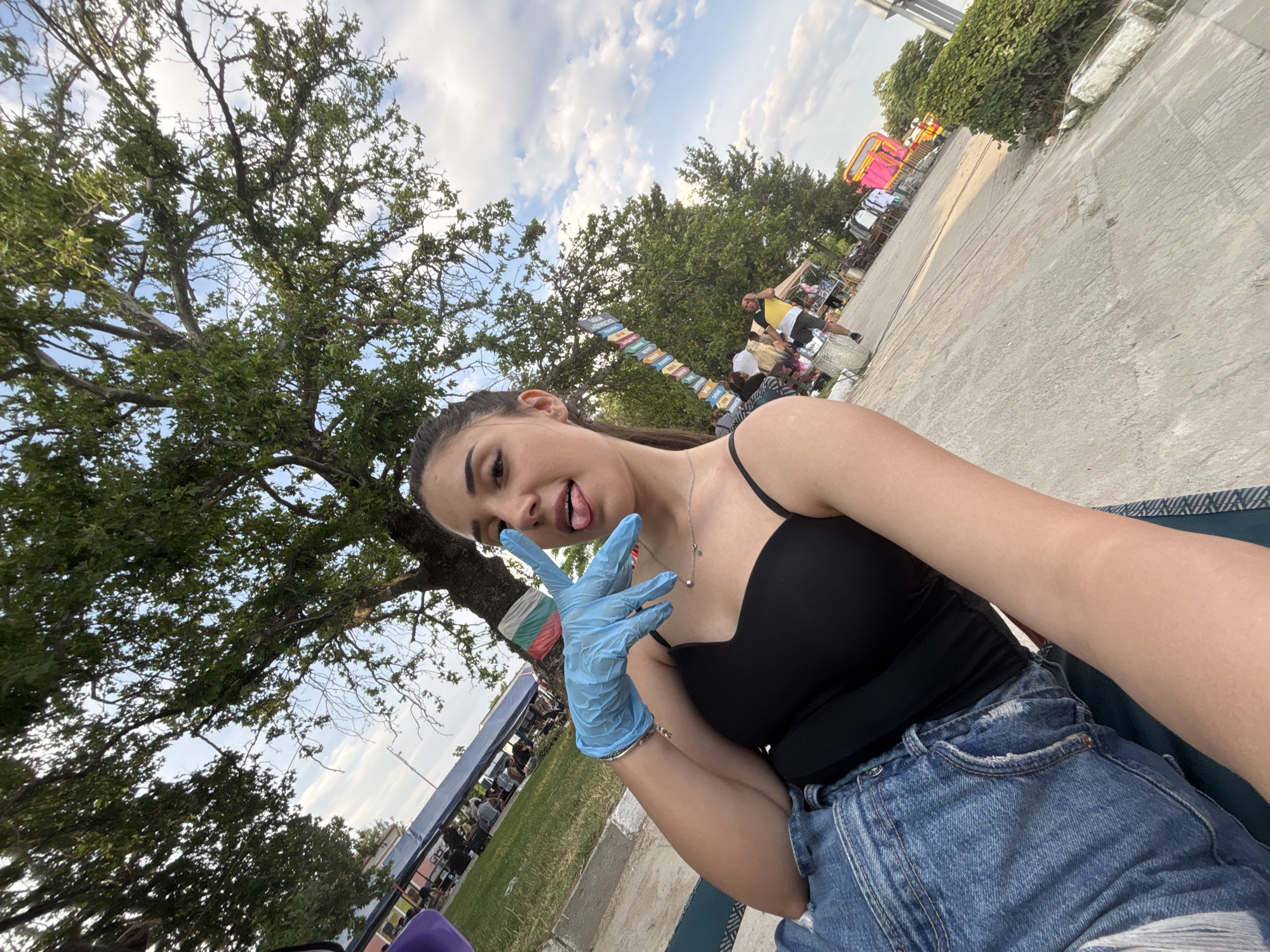 The image depicts a young woman taking a selfie in an outdoor public setting, identified as Kostievo, Bulgaria, by a visible Bulgarian flag. The woman, with dark hair pulled back, is seen from the chest up, angled slightly to the left. She is wearing a black spaghetti-strap top and blue denim jeans, partially visible. On her right hand, she wears a light blue nitrile-style glove and forms a peace sign (V-gesture) with her index and middle fingers, extending towards the camera. Her tongue is slightly protruded. She has a silver necklace with a small pendant and a silver bracelet on her right wrist.
She appears to be sitting or reclining on a dark green patterned fabric, laid on a concrete path or sidewalk. To her left, a grassy area hosts numerous people gathered under blue and white canopies or tents, suggesting a market, fair, or public event. Large trees with green foliage dominate the upper left background and extend across the top of the image.
To her right, a thick tree trunk is visible, around which a Bulgarian flag (white, green, red horizontal stripes) is tied. Further along the concrete path, a vertical structure composed of multiple colorful, rectangular signs is visible. Several individuals are present on the path, including a person in a yellow and black shirt. Stalls displaying various goods, including clothing, are set up along the path, with a bright pink structure visible in the far right midground. The sky above is partly cloudy with patches of blue visible through the tree canopy.