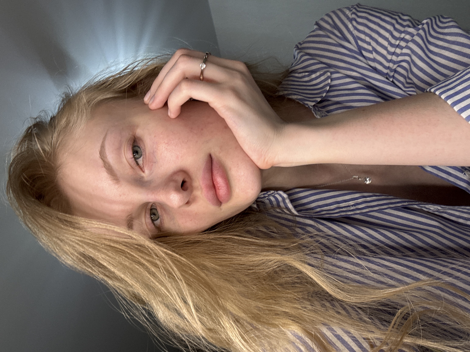 A young, fair-skinned woman with long blonde hair is reclining against a light gray wall. She is wearing a blue and white striped collared shirt, and a silver ring is visible on her left hand. She appears to be resting her face against her hand. A silver necklace is visible at her collarbone. The lighting is soft and diffused. The location of the photograph is unspecified and cannot be discerned from the image.