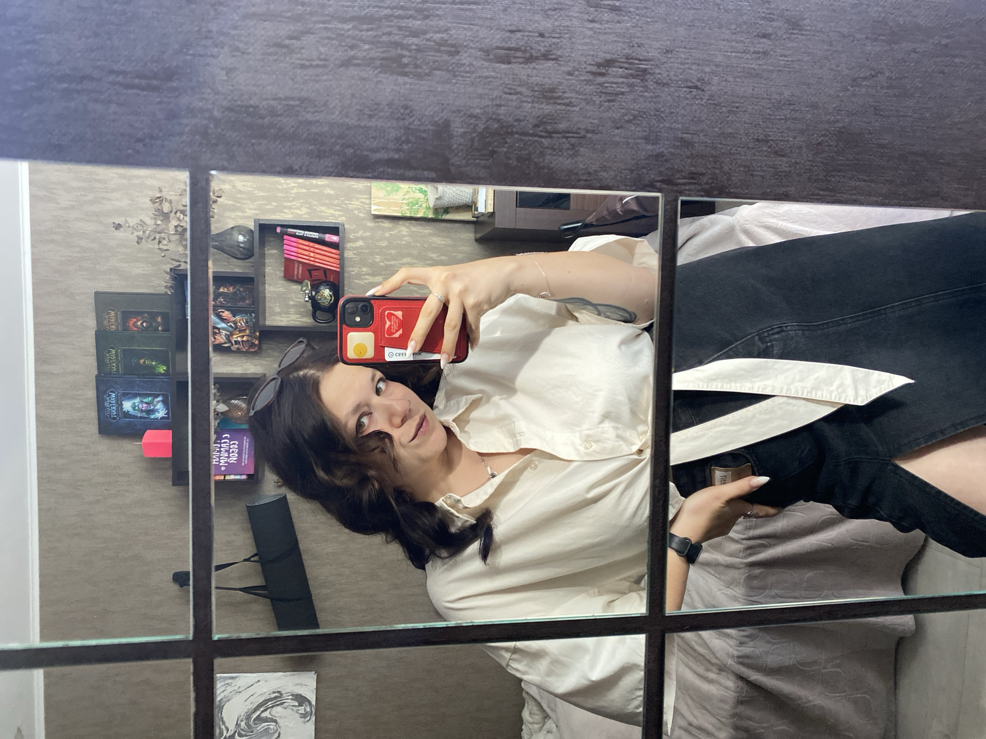 A young woman with dark, wavy hair is reflected in a large mirror, taking a selfie with her red smartphone. She is wearing a loose, cream-colored button-down shirt tied at the waist and a dark denim skirt with a high slit. The mirror is framed by dark brown wood and has a grid pattern that divides the reflection into sections. In the background, visible in the mirror, are shelves with books, a pink candle, a small vase, and a framed picture on the wall. A bed with a textured grey comforter is also visible. The room has beige wallpaper. The image appears to be taken indoors, likely in a residential setting. The person's attire suggests a casual or semi-casual style. There are no indications of the specific city of Chaikovsky, Russia within the image itself.