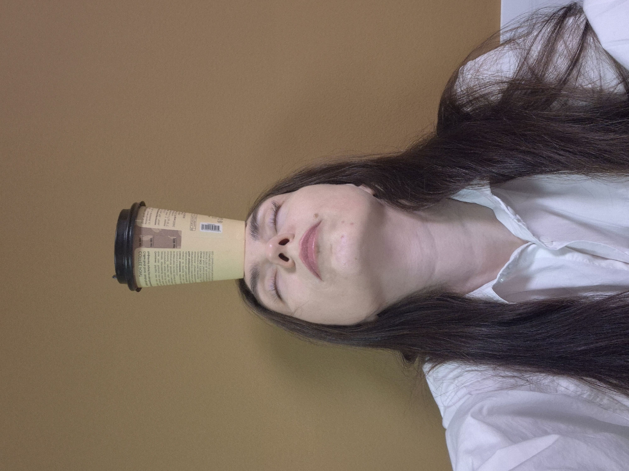 The image shows a woman lying down on a brown surface, possibly a table or floor. She is wearing a white shirt and has long, dark hair. A disposable coffee cup is balanced on her forehead. The background is a plain brown wall. The location appears to be an indoor setting.