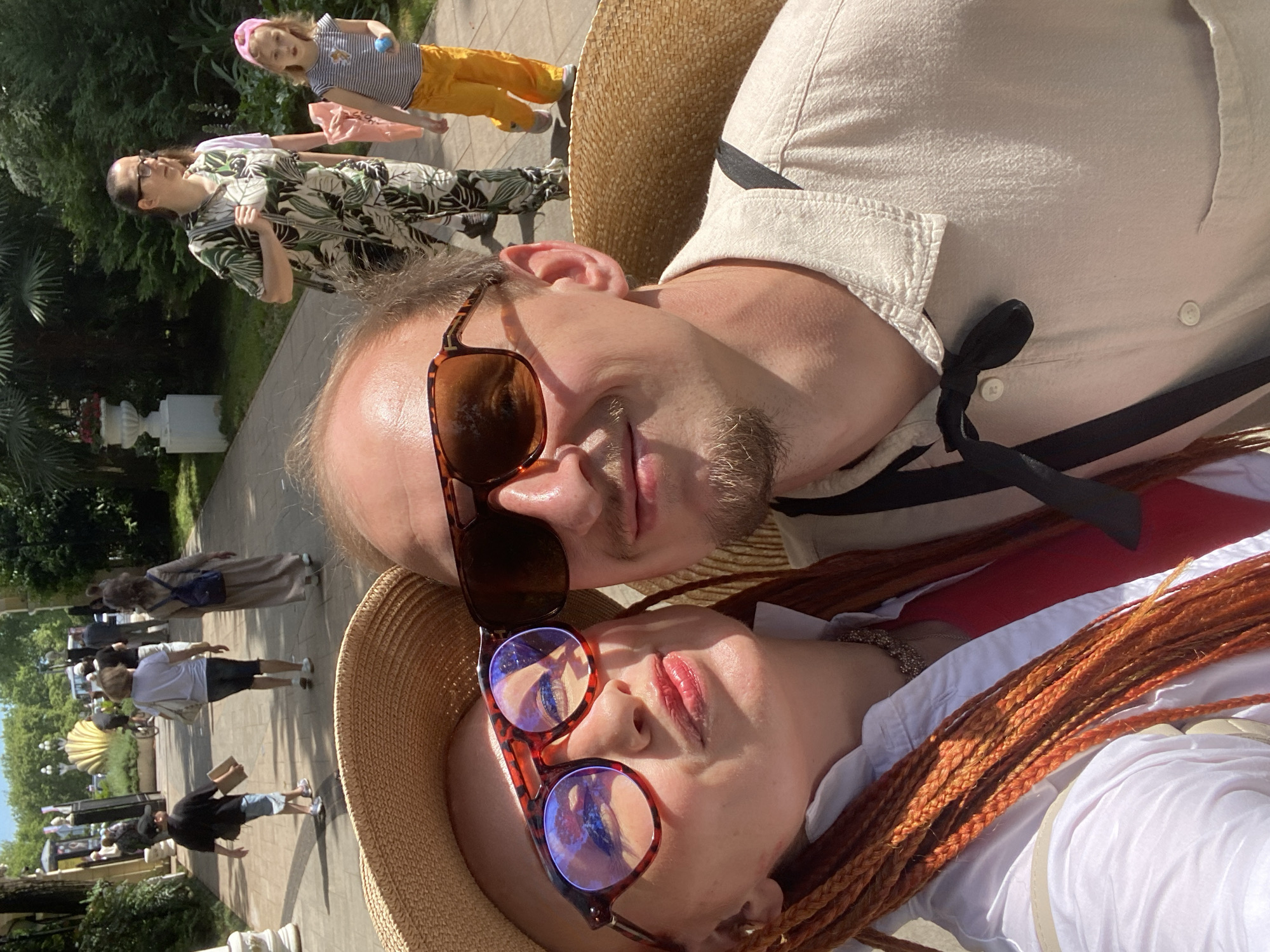 The image depicts a selfie taken in an outdoor urban park-like setting, possibly in Sochi, Russia, given the presence of palm trees and lush greenery. In the foreground, a man and a woman are close to the camera, both wearing wide-brimmed straw hats and sunglasses. The woman, on the left, has distinctive orange braided hair and is wearing a white collared shirt over a red top, with a glittering necklace. The man, on the right, has short facial hair and is wearing a light-colored collared shirt with a black ribbon tied at the collar.
In the mid-ground, several other people are walking along a paved pathway. On the left, a young boy in a black t-shirt and a baseball cap is walking with a book or folder. Further down the path, a woman in a white t-shirt and dark shorts is walking away from the camera, carrying a bag. Beside her, a woman in a long, flowing dress with a blue backpack is also walking away. In the background, a woman in a patterned green and white outfit is walking with a child wearing a pink headband and yellow pants. The environment appears sunny with dappled shadows cast by the trees, and there are some decorative planters and architectural elements visible.