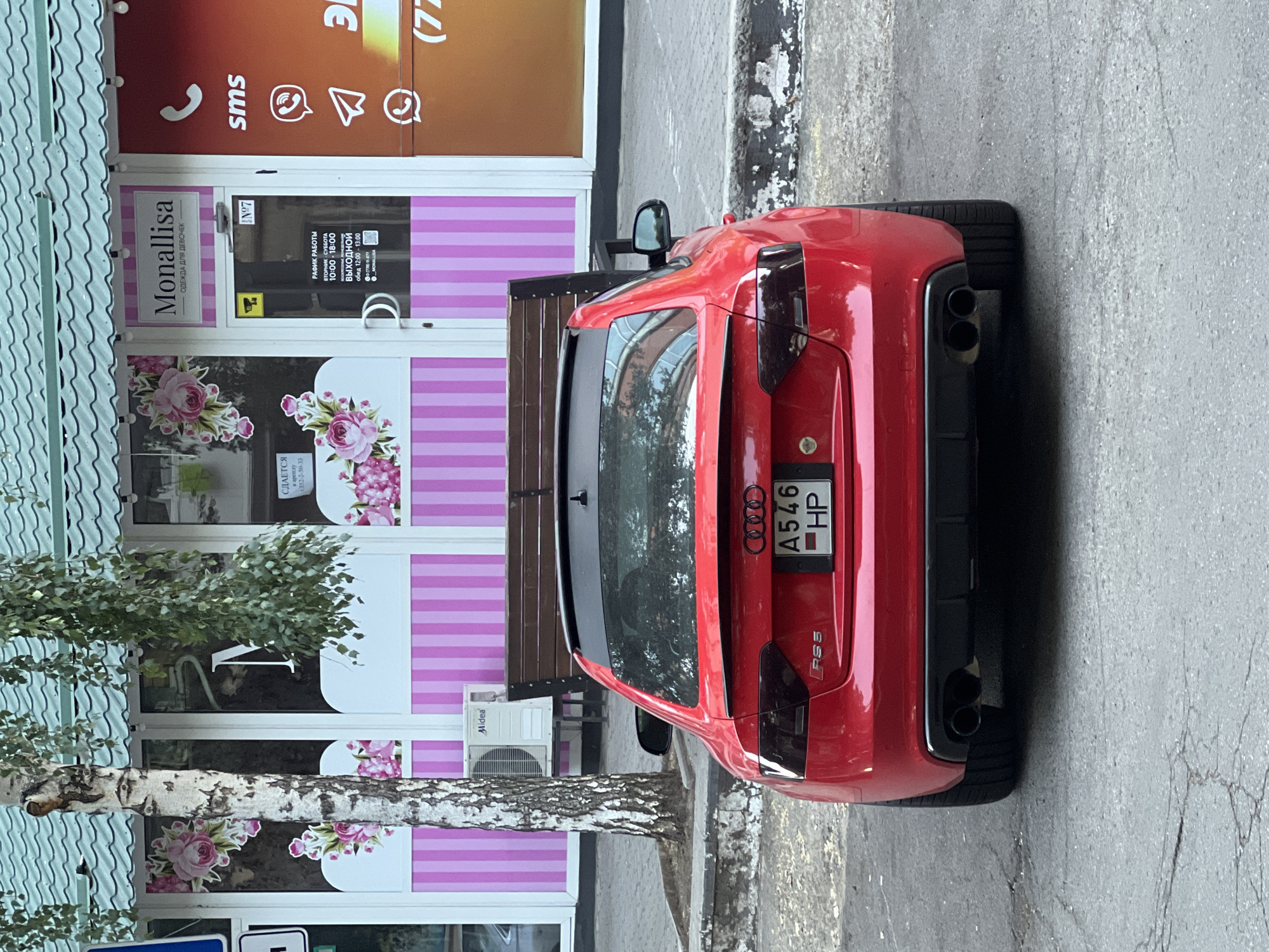 The rear view of a red Audi RS5 is parked on a paved street in front of a storefront. The storefront has a green roof and a pink and white striped facade with floral decorations. A sign above the door reads "Monallisa" with the tagline "ОДЕЖДА ДЛЯ ДЕВОЧЕК" (Clothing for girls). Below the sign, a schedule indicates business hours. A tree with a white trunk is to the left of the storefront, and a wooden bench is positioned in front of the store. The car's license plate is visible and reads "A546 HP". The overall scene appears to be in an urban environment.