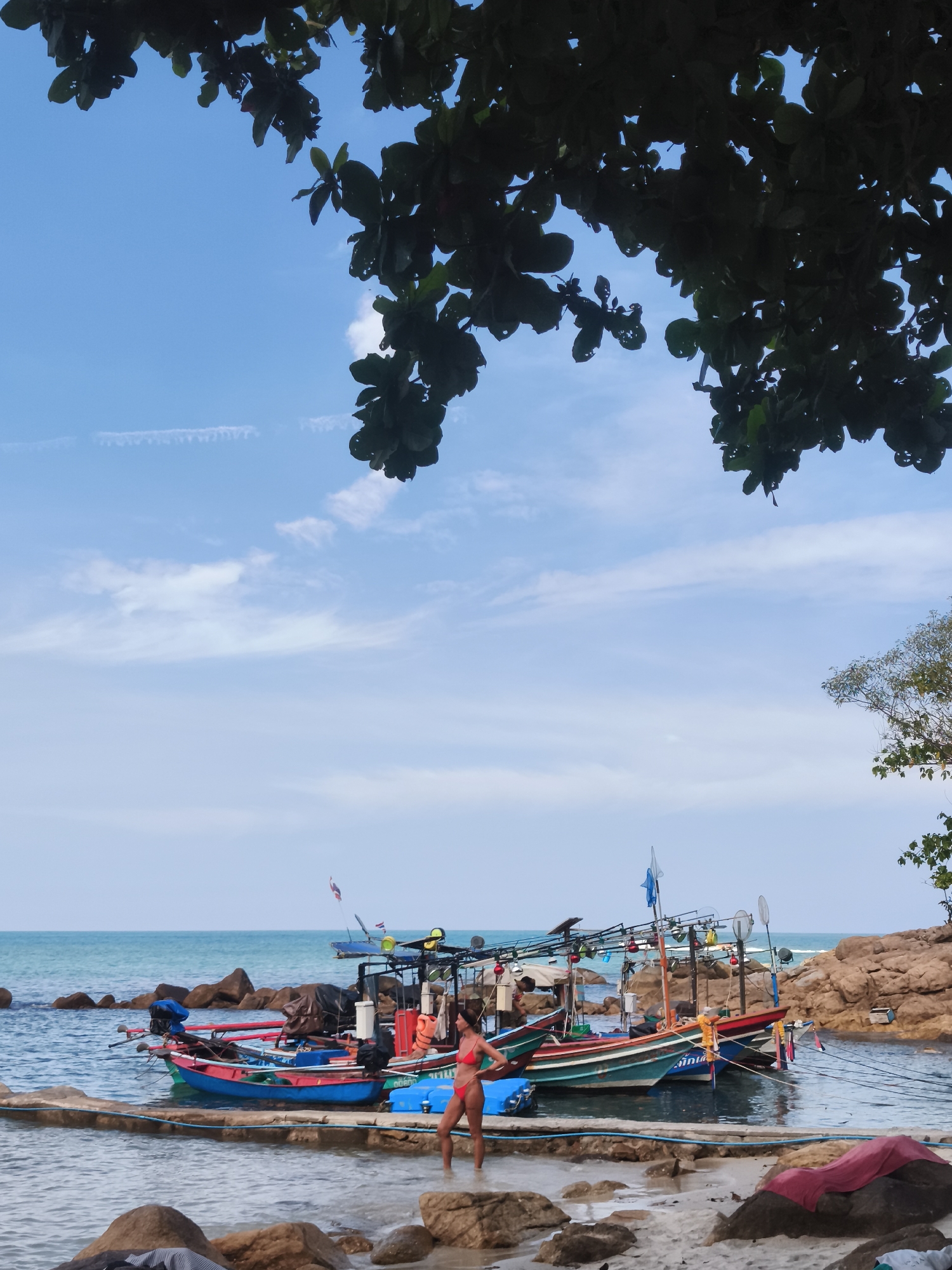 A woman in a red bikini stands in shallow water near a concrete barrier. She is facing a row of colorful fishing boats docked in a sheltered cove. The boats, painted in shades of blue, green, and red, are equipped with outriggers and various fishing gear. Behind the boats, rocky outcrops and vegetation line the shore. The sky above is a clear blue with scattered white clouds, and the leaves of a tree hang into the frame from the top. The scene appears to be a beach in the Ko Samui District of Thailand.