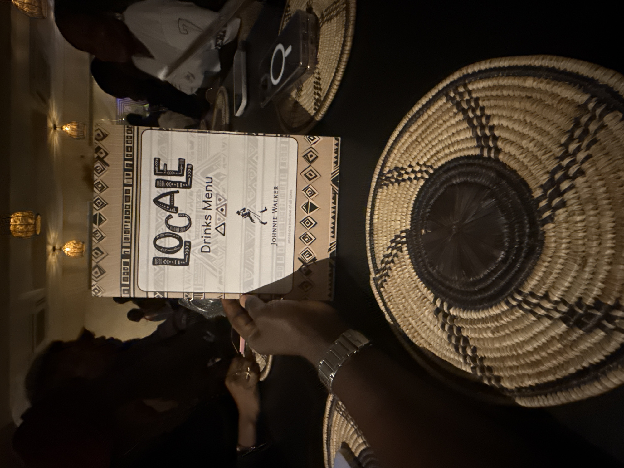 The image captures a table setting in Locale Ikoyi, Lagos, Nigeria. A menu titled "LOCALE Drinks Menu" is prominently displayed. Beside the menu are two woven baskets with intricate black and white patterns. A person's hand with a silver wristwatch is visible, seemingly holding or touching the menu. A phone lies on a smaller woven basket to the top left. Wall lights can be seen in the background. Additional people are present, but their features are not distinct.