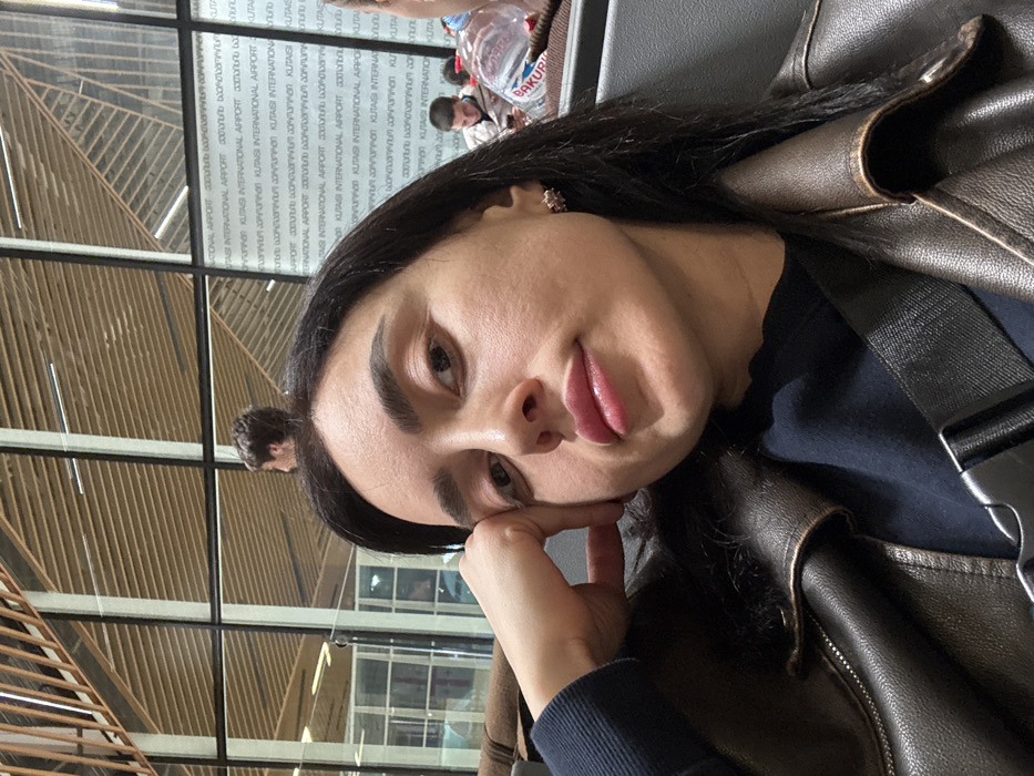 A woman poses for a selfie in an airport setting. She's resting her head on her hand with a relaxed expression. She's wearing a black top and a brown leather jacket with a seatbelt or bag strap visible. In the background, a window displays text repeating "KUTAISI INTERNATIONAL AIRPORT" in various languages. Other passengers and items like a water bottle can be seen through the window, suggesting a waiting area.