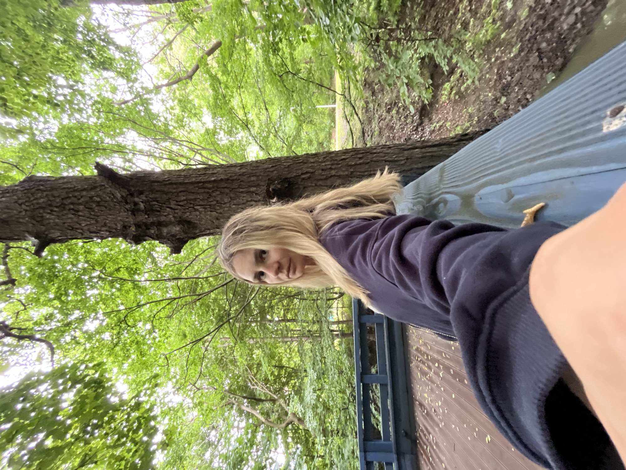 An outdoor, medium shot captures a blonde woman standing on a wooden deck. She is positioned to the right of the frame, facing the camera with a neutral expression. She has on a dark blue hooded sweatshirt and light blue pants. The deck has a wooden railing, painted blue, that extends across the foreground. Behind the railing, a wooded area with lush green trees creates a natural backdrop. The lighting suggests it is daytime, possibly overcast due to the diffused light filtering through the leaves. The image does not provide specific information about a location in the city of Pionersky, Russia.