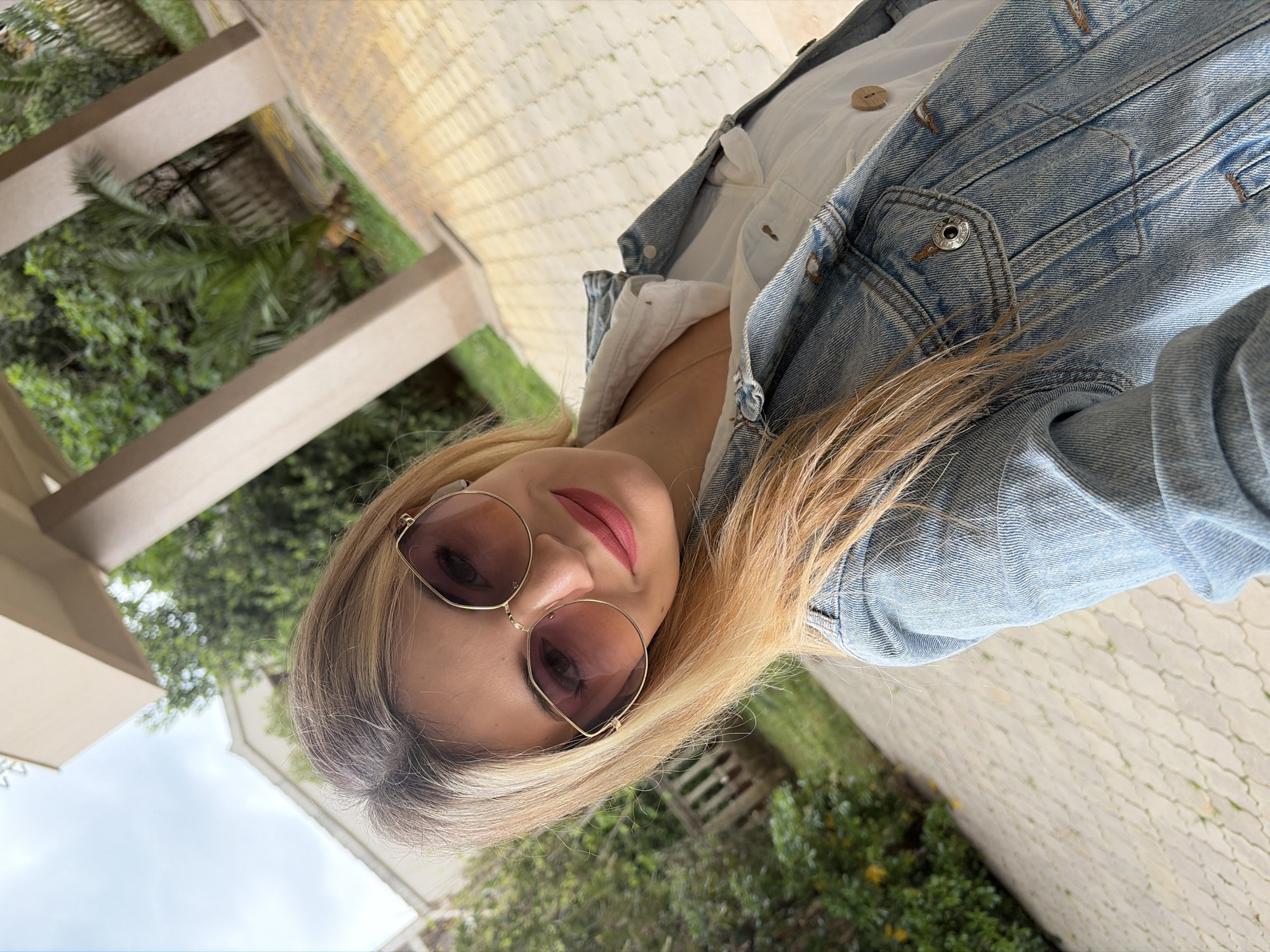 A woman with blonde hair and sunglasses is the main subject of the image. She is wearing a white shirt and a denim jacket. The background appears to be an outdoor area with plants and possibly some architectural structures. While it is impossible to confirm the location is Budva, Montenegro without additional information, the presence of lush greenery and what seems like a patio or walkway suggest an outdoor setting that aligns with the landscape of Budva.