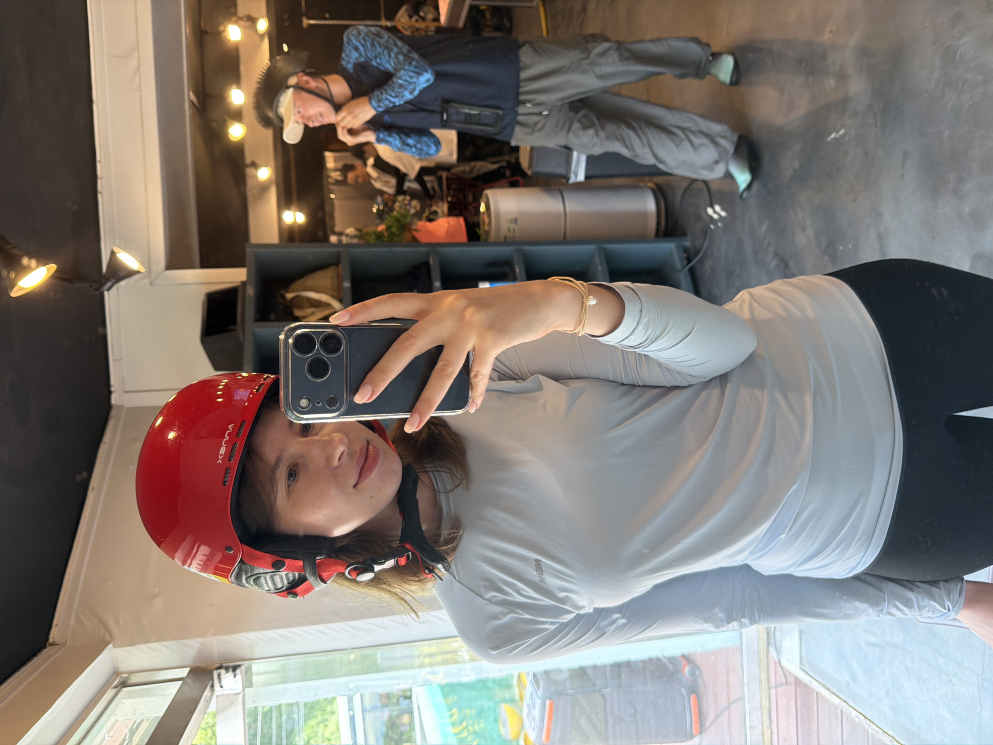 A young woman with brown hair and light skin, wearing a red helmet and a light gray long-sleeved shirt, takes a mirror selfie with her phone. She is standing in what appears to be a retail store with a large window on the left side. In the background, a man wearing a dark blue long-sleeved shirt with a blue pattern, gray cargo pants, and a visor is adjusting his helmet. The store has shelves filled with various items, including clothing and accessories. The floor is polished concrete, and there are several lights mounted on the ceiling. The overall impression is of a casual, indoor environment, possibly a surf shop or an outdoor gear store. The location is identified as Seo-myeon, South Korea.