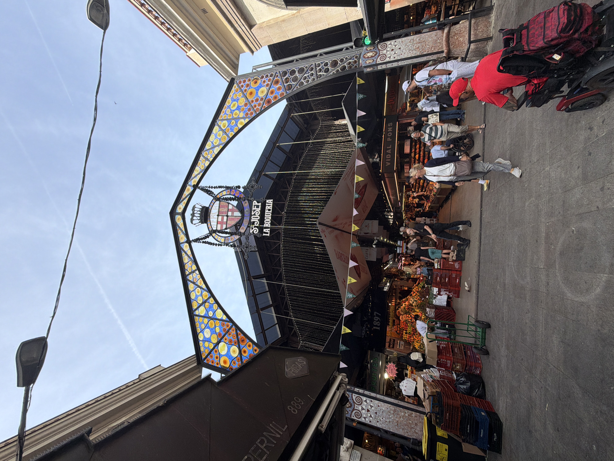 The image shows an outdoor market scene under a grand, ornate archway. The arch is decorated with stained glass in geometric patterns of blue, yellow, and orange. Above the arch, a crest with the coat of arms of Barcelona is visible, with the text "ST. JOSEP LA BOQUERIA" underneath. The market stalls are bustling with activity. In the foreground, a person in a red shirt is seated in an electric wheelchair. Other people are walking through the market, some carrying bags or pushing carts. Stalls are filled with various goods, including a large display of fruits. The ground is paved, and buildings with architectural details line the sides of the market. The sky above is blue with some wispy clouds and a contrail from an aircraft. The overall impression is a lively and vibrant marketplace. Based on the signage "ST. JOSEP LA BOQUERIA", this market is La Boqueria Market, located in Barcelona, Spain, not L'Hospitalet de Llobregat.