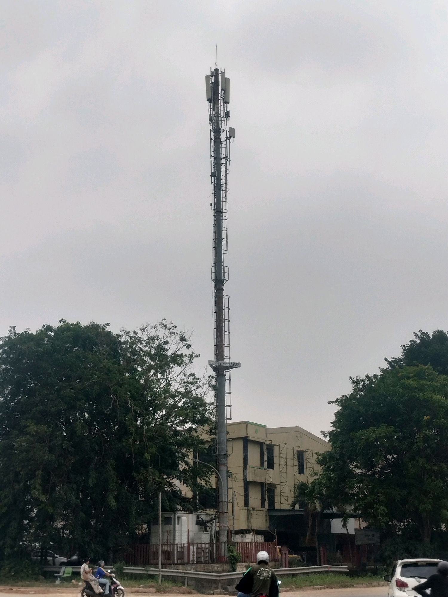 The image depicts a tall telecommunications tower positioned centrally in the frame, reaching towards an overcast sky. The tower is constructed from metal lattice work and is adorned with various antennas and equipment at its upper sections. To the left and right of the tower, large, verdant trees dominate the middle ground, their foliage dense and dark green. Behind the trees and to the right of the tower, a light-colored, multi-story building with several windows is visible. The building appears to be industrial or commercial in nature, with some scaffolding visible on its facade. In the foreground, a paved road runs horizontally across the bottom of the image. On this road, a white car is stationary or moving slowly on the right side. Two individuals are on a motorbike in the left portion of the foreground, with one person driving and another as a passenger. The environment suggests an urban or semi-urban setting, likely in a tropical climate due to the lush vegetation. The sky is a uniform light gray, indicating cloudy or possibly rainy weather conditions.