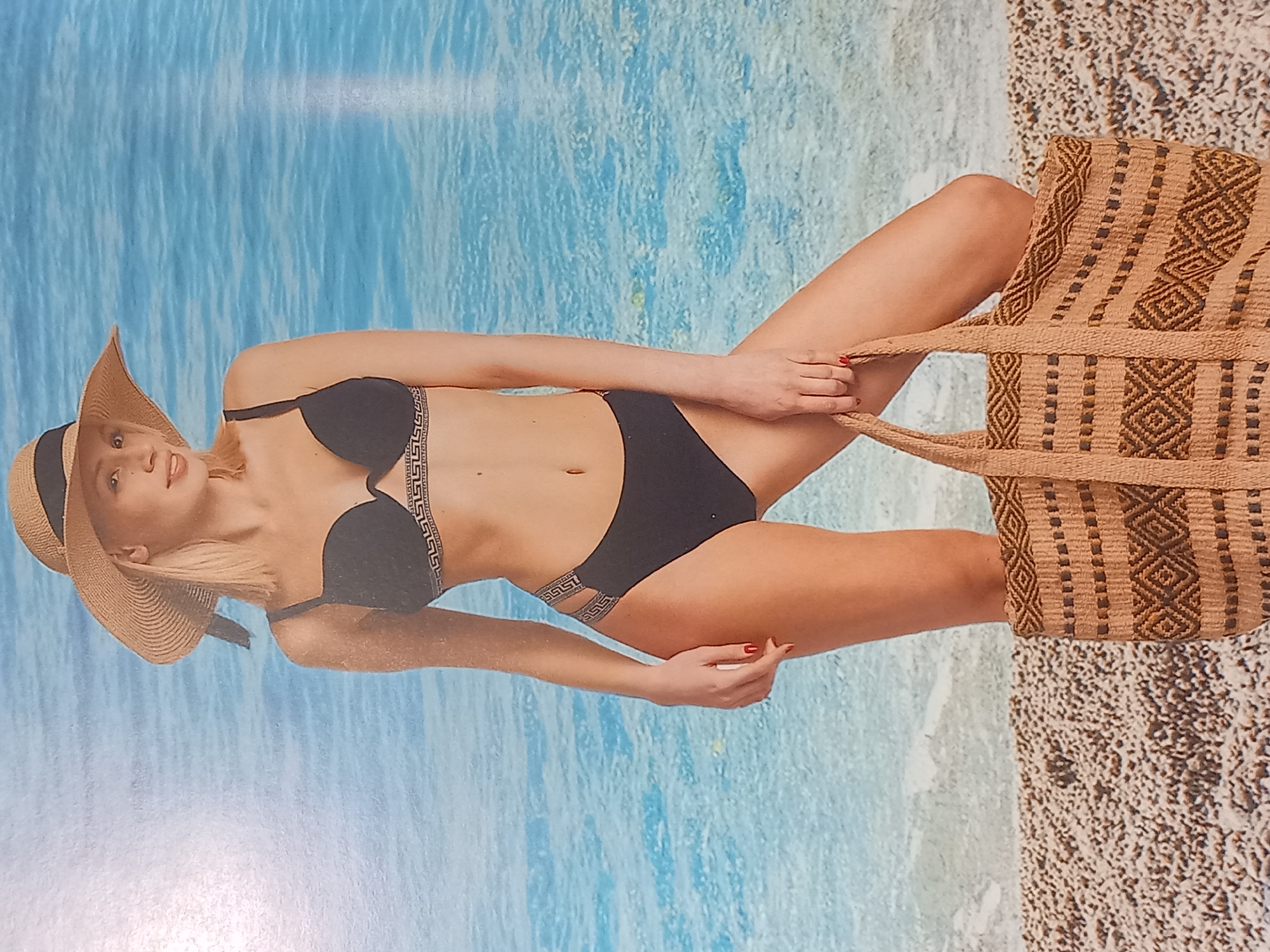 A blonde woman stands on a pebble beach, holding a woven tote bag in her right hand. She is wearing a black bikini with a Greek key pattern trim on the bikini top and a decorative strap detail on the bikini bottoms. A wide-brimmed straw hat with a black ribbon adorns her head. The background depicts a blurred image of the ocean under a clear sky. The woman is posed looking to her right, with her left hand resting on her left hip. Her fingernails are painted red. The lighting suggests daytime.