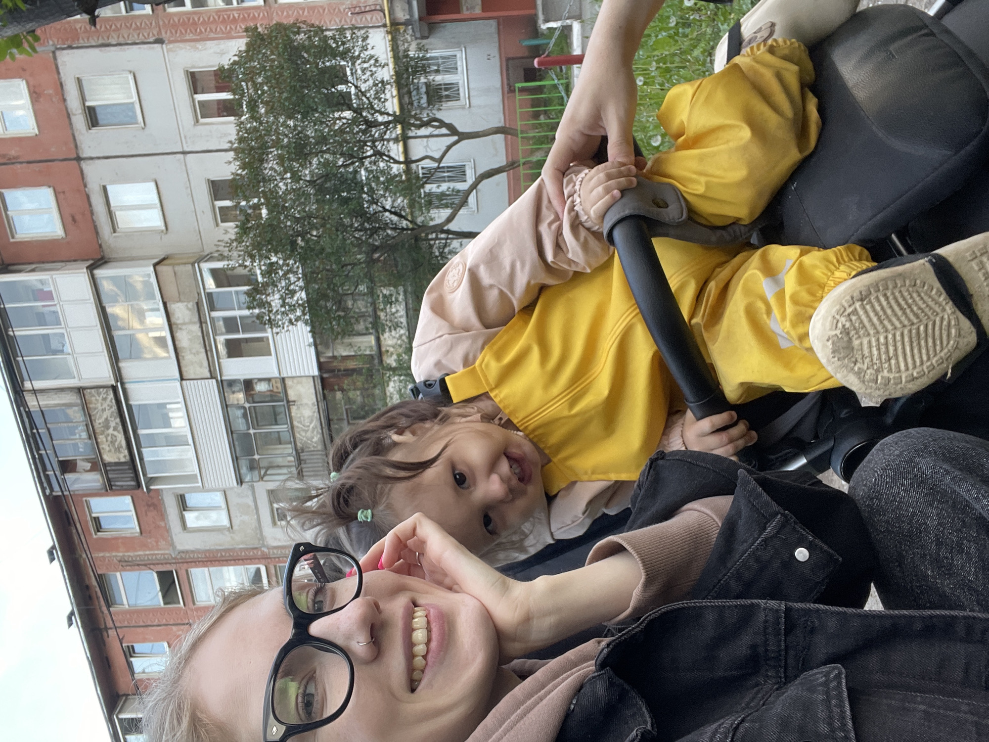 The image captures a selfie taken by a fair-skinned woman with blonde hair and wearing black-rimmed glasses. She is smiling. She has a nose ring on her left nostril. In the background, a young child with brown hair and wearing a yellow and beige jacket is in a stroller. The child also appears to be smiling. In the background, there is an apartment building. According to metadata, the image was taken in Izhevsk, Russia. A playground is present behind the apartment building.