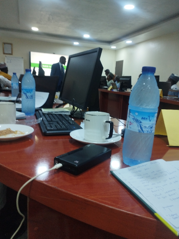 The image shows a table in a meeting room at 10.929, 11.476 in Ashaka, Nigeria, with a computer, a laptop, a power bank, a coffee cup, two bottles of water, a plate, a notebook, and a few other objects on it. There are several people, including a man standing in the background, and several people sitting behind the table. There are fluorescent lights in the ceiling. There is a whiteboard in the background, with a whiteboard marker sitting on it. The room has a white ceiling and white walls.