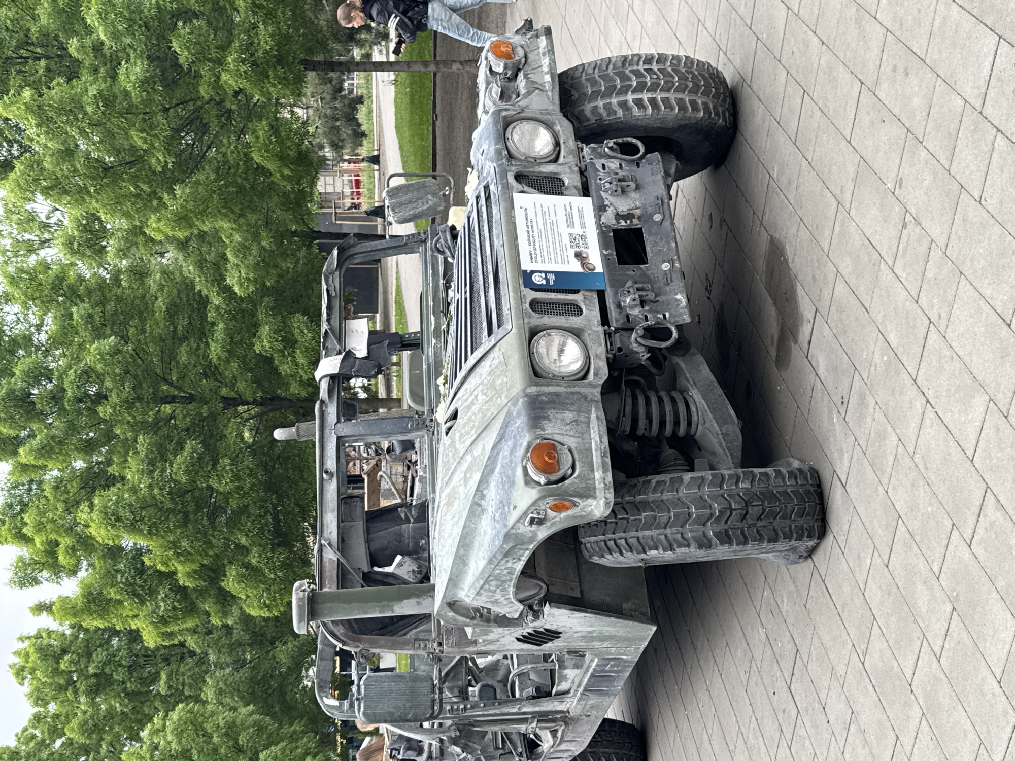 A damaged Humvee is parked on a paved surface, likely a sidewalk or pedestrian area, in Kyiv, Ukraine. The vehicle is significantly distressed, with visible wear and tear indicative of possible combat damage. A sign with text and QR codes is attached to the front grille, presumably providing information or context about the vehicle. Several trees provide a backdrop to the scene, suggesting a public space or park. A person can be seen behind and adjacent to the vehicle appearing to take photographs.