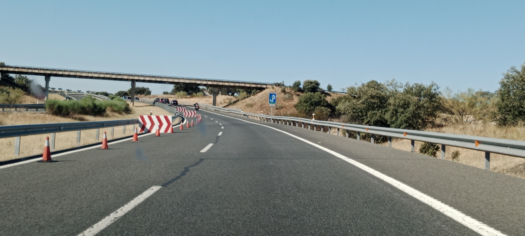 The image depicts a highway scene with a clear blue sky. In the foreground, a grey asphalt road with white lane markings curves to the right. Traffic cones and chevron signs are placed along the right side of the road, indicating a lane closure or diversion. A silver guardrail runs parallel to the road. In the middle ground, a concrete overpass spans across the highway. Further back, several cars and a motorcycle are visible on the road, moving away from the viewer. The landscape is characterized by dry, yellowish grass and scattered green trees on a sloping hill. A blue parking sign with a white 'P' is visible on the right side of the road. The scene appears to be on a highway near Jaraicejo, Spain.