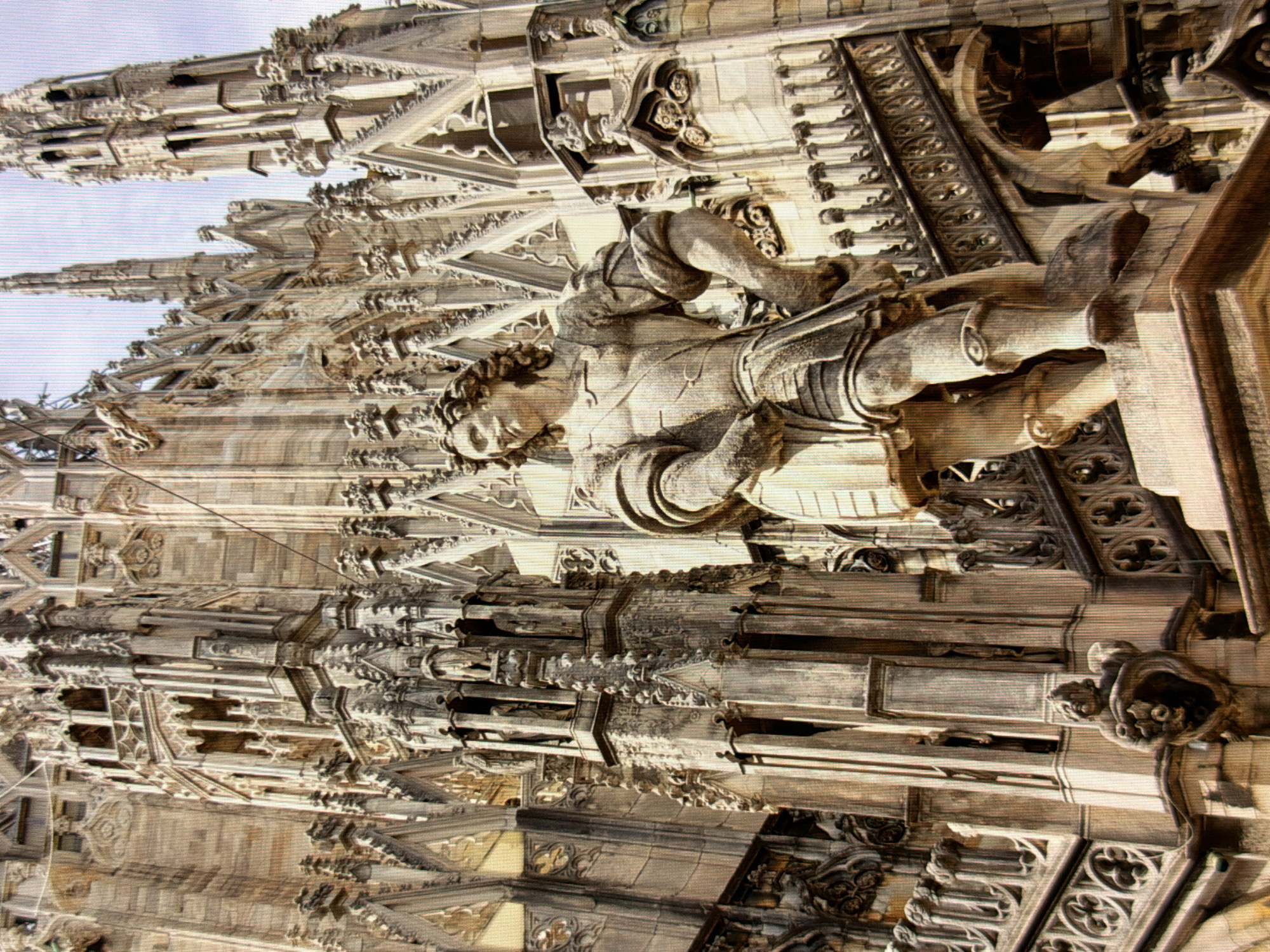 The image depicts a close-up, low-angle view of a statue of a male figure in ornate armor, standing against the backdrop of a large Gothic-style building. The statue is made of stone and is detailed, showing muscular anatomy, a mustache, and curly hair. The building behind it is characterized by intricate stonework, spires, arched windows, and numerous small statues and decorative elements, indicative of Gothic architecture. The sky above is a pale blue. The scene is a detailed architectural study of a historic structure and its sculptural embellishments. The location is not Kyiv, Ukraine, but rather Milan Cathedral, Italy.
