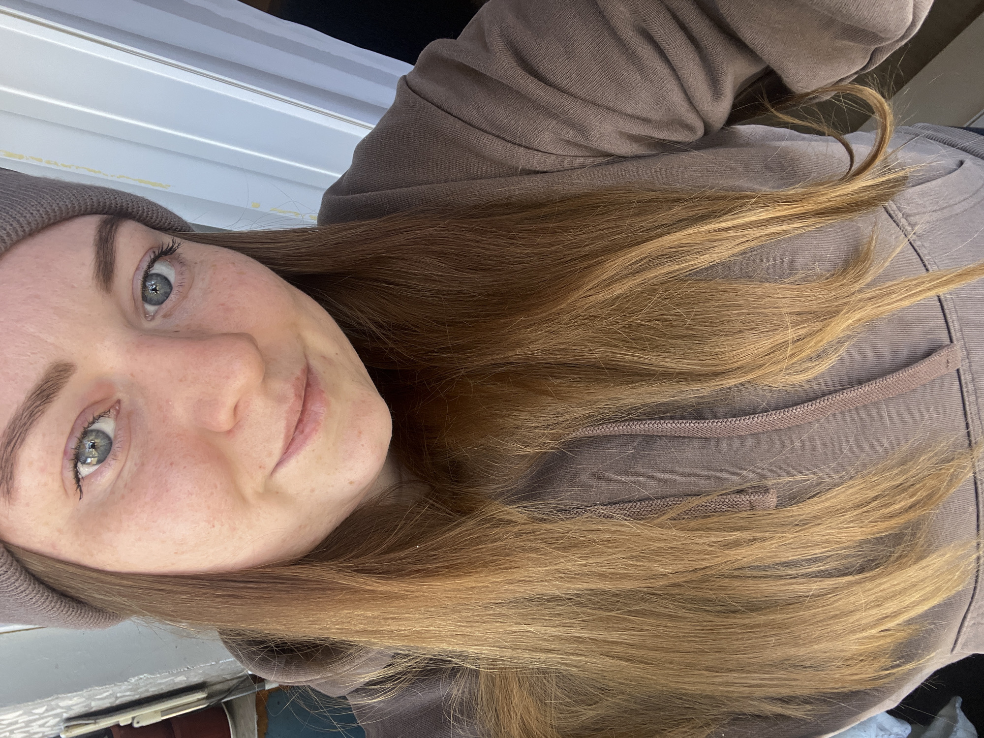 A young woman with fair skin, freckles, and light brown hair is wearing a brown beanie and hoodie. Her eyes are light blue-grey. The background shows a white window frame and a hint of an interior space. No elements specifically indicate the location of Раздолье, Russia.