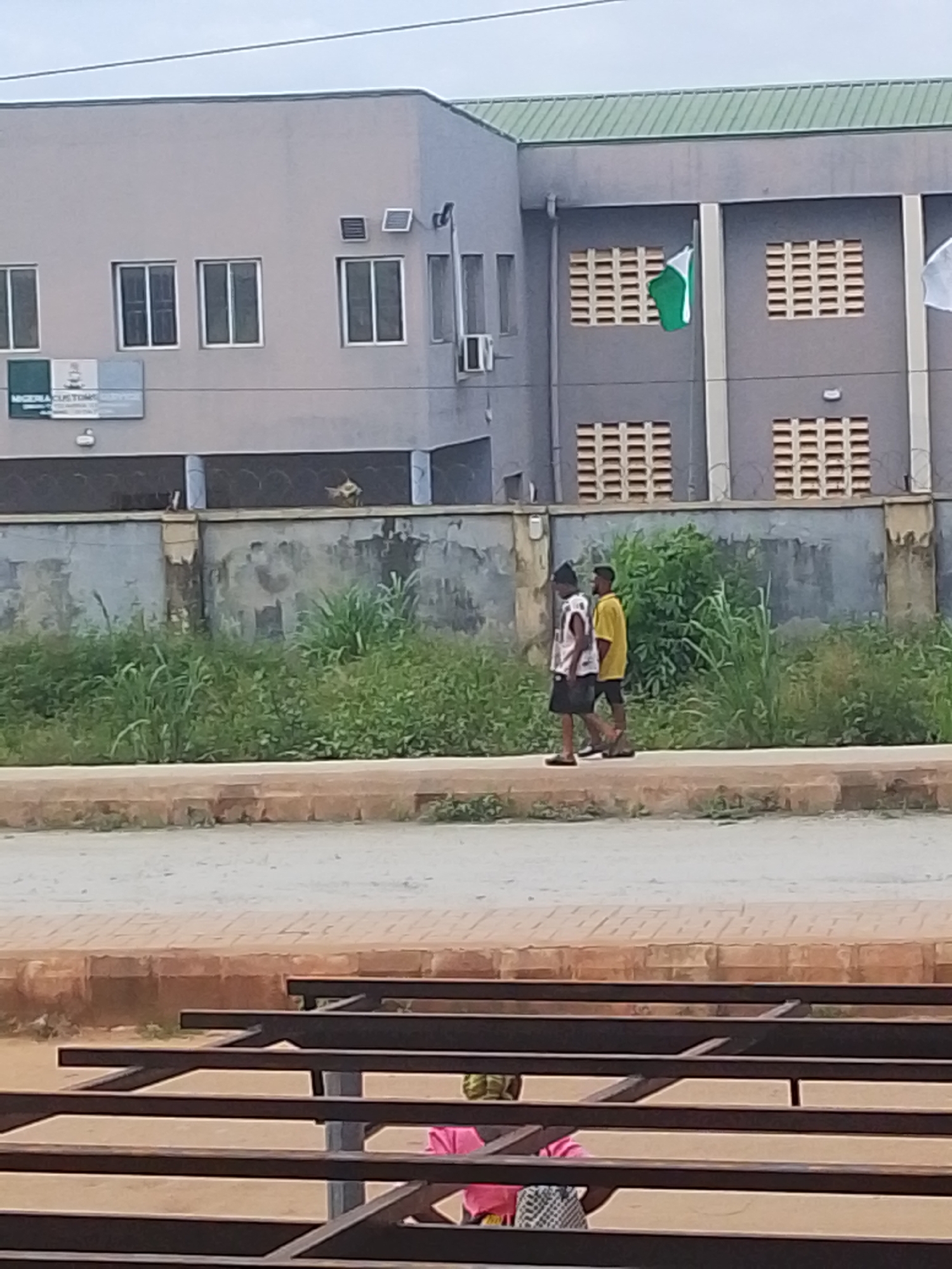 The image captures a street scene in Abeere, Nigeria, with a building prominently featured in the background. The building is a multi-story structure with a gray facade and a green roof. It has several windows with white frames and louvers, as well as what appear to be air conditioning units. A Nigerian flag is displayed on a flagpole attached to the building. In front of the building is a grey concrete wall topped with barbed wire. A section of the wall appears to be adorned with graffiti. Two young men are walking on a paved sidewalk in front of the wall. One man, on the left, wears a white tank top and black shorts, with a dark head covering. The other man, on the right, is wearing a yellow t-shirt and dark shorts. They are walking away from the camera. Behind the sidewalk and wall, there is dense green vegetation. The foreground of the image shows a section of a brown brick pathway and dark metal structures, possibly part of a bench or seating area. The sky is overcast.