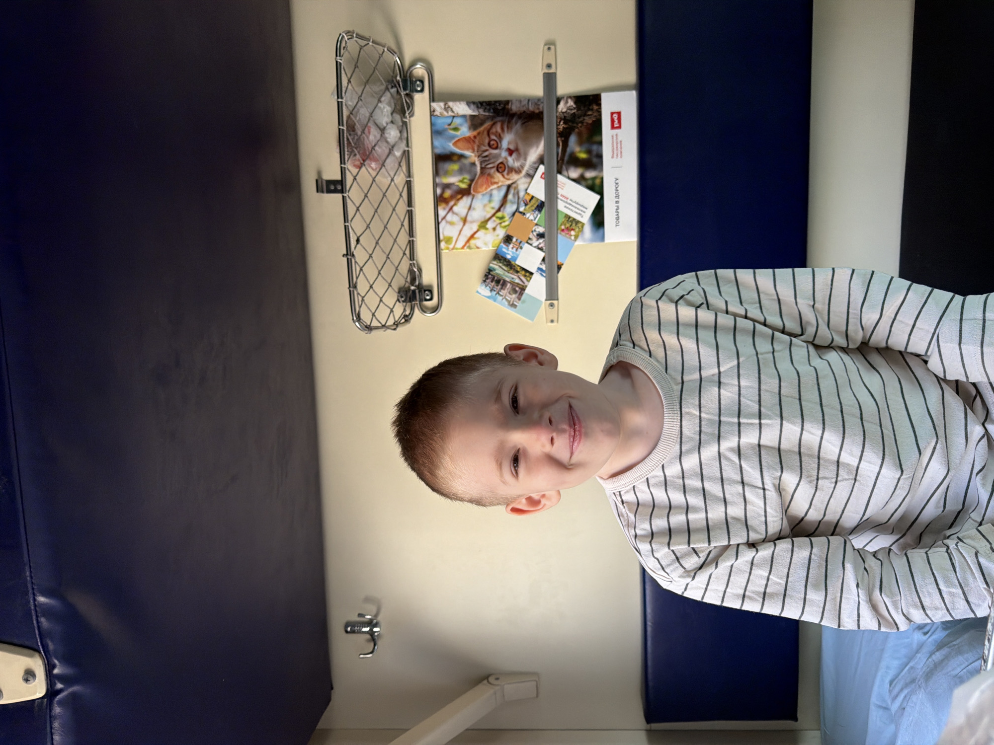 A young boy with short, light-brown hair and fair skin is smiling at the camera. He is wearing a striped, long-sleeved shirt. The boy is in what appears to be a train compartment. Behind him are a blue padded seat, a white wall, and overhead storage. A metal mesh basket is mounted on the wall. There is a magazine or brochure with a cat picture, along with postcards. A small metal hook and a folding table are also visible. Without additional information or reference points in the image, determining the location in Krasnodar, Russia, is impossible.