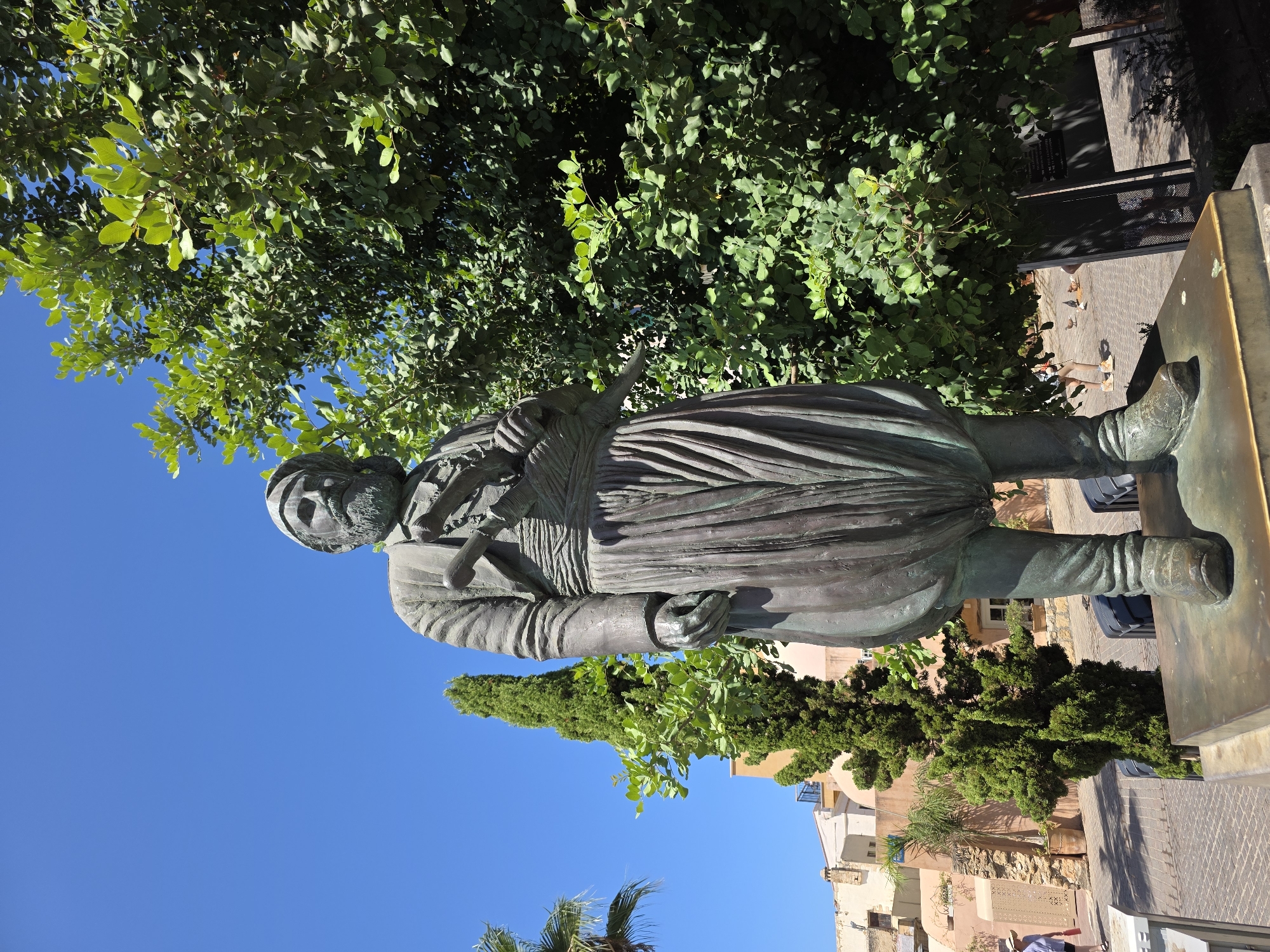 A full-body bronze statue of a man in traditional Cretan attire stands in a public space in Chania, Greece. The man is depicted with a full beard, wearing a vest over a tunic, a sash around his waist, and baggy trousers tucked into boots. He holds a sword or dagger in his right hand, which is tucked into his sash. The statue is positioned on a pedestal, with lush green trees and a clear blue sky in the background. To the left of the statue, a tall cypress tree stands, and behind it, buildings with light-colored facades can be seen. In the background, a woman wearing a straw hat and a light-colored shirt walks away from the viewer on a cobblestone path, accompanied by pigeons. The overall atmosphere suggests a warm, sunny day in an outdoor urban setting.