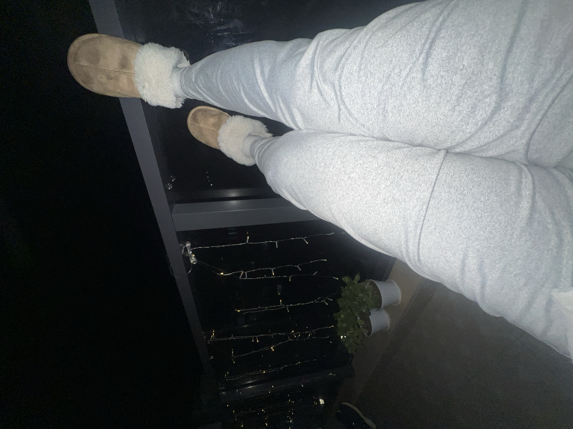 The image displays a nighttime scene from a balcony in the city of Kiryat Bialik, Israel. The lower legs and feet of an individual are visible in the foreground, extended horizontally into the frame. The individual is wearing light grey athletic pants and tan, fur-lined slippers. Their feet are positioned above a dark-colored balcony railing, from which several strands of illuminated, warm white LED string lights are suspended vertically. Below the railing, on the balcony surface, two green potted plants are present in white cylindrical containers. The background consists of a dark night sky with distant, scattered city lights, indicating an elevated vantage point.