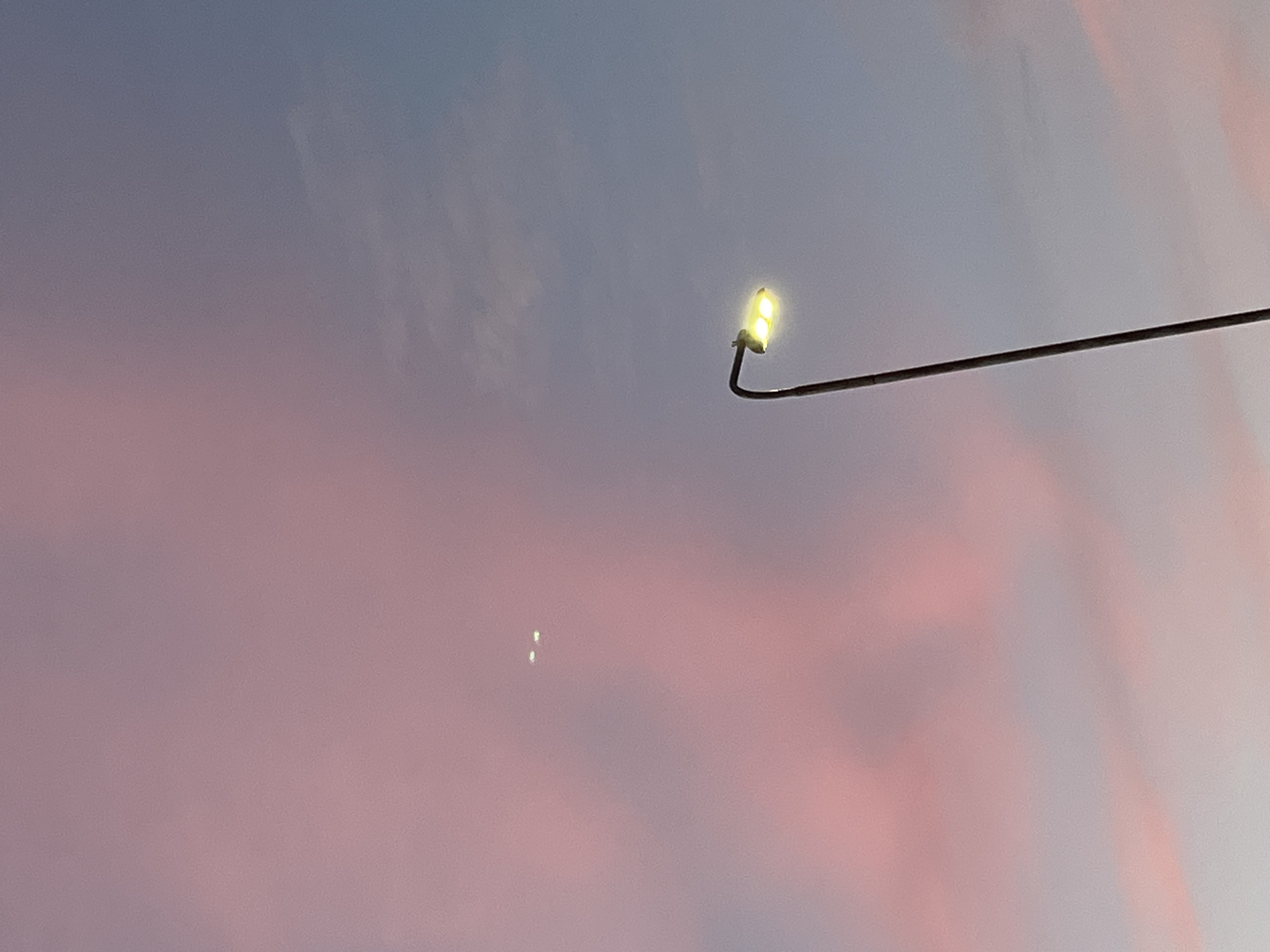 This is a vertical, low-angle, medium shot of a street lamp against a twilight sky. The sky is a gradient of soft pinks and blues, with some wispy clouds. The street lamp is dark metal, with two yellow lights illuminated at the top. In the upper-left quadrant of the image, two small, bright points of light are visible, possibly distant lights or celestial objects. The overall mood is serene and somewhat mysterious. The image was taken in Saint Petersburg, Russia.