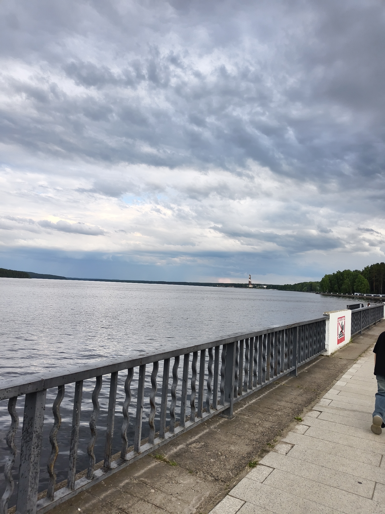 The image shows a view of the Kama River from a promenade in Chaikovsky, Russia. A grey metal railing runs along the edge of the promenade, separating it from the river. The river is calm with a rippled surface. In the distance, a lighthouse is visible on the far bank, which is lined with a dense forest. The sky is overcast with dramatic grey clouds. A person, only their lower legs and feet are visible, is walking away from the camera on the paved promenade. There is a white sign with red lettering on the railing.