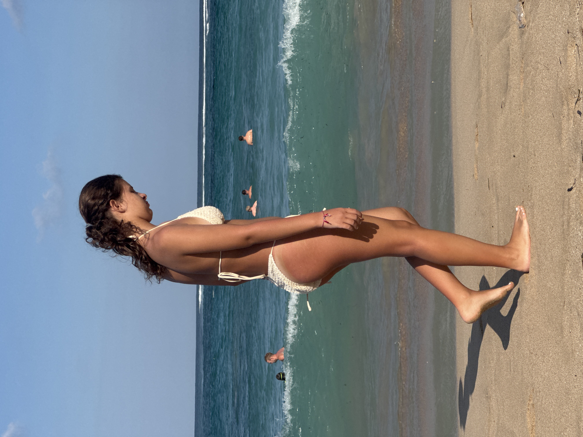 In Kuta Selatan, Indonesia, a young woman with dark, curly hair pulled back walks along a sandy beach towards the right of the frame. She is wearing a cream-colored crochet bikini. Her body is angled in profile, with her head bowed slightly. The ocean is visible behind her, with turquoise waves breaking and a white foam line stretching across the horizon. Several people are in the water, appearing as small figures in the distance. The sky is a clear, bright blue. The sand underfoot shows footprints and small debris. A shadow of the woman is cast on the sand to her left.