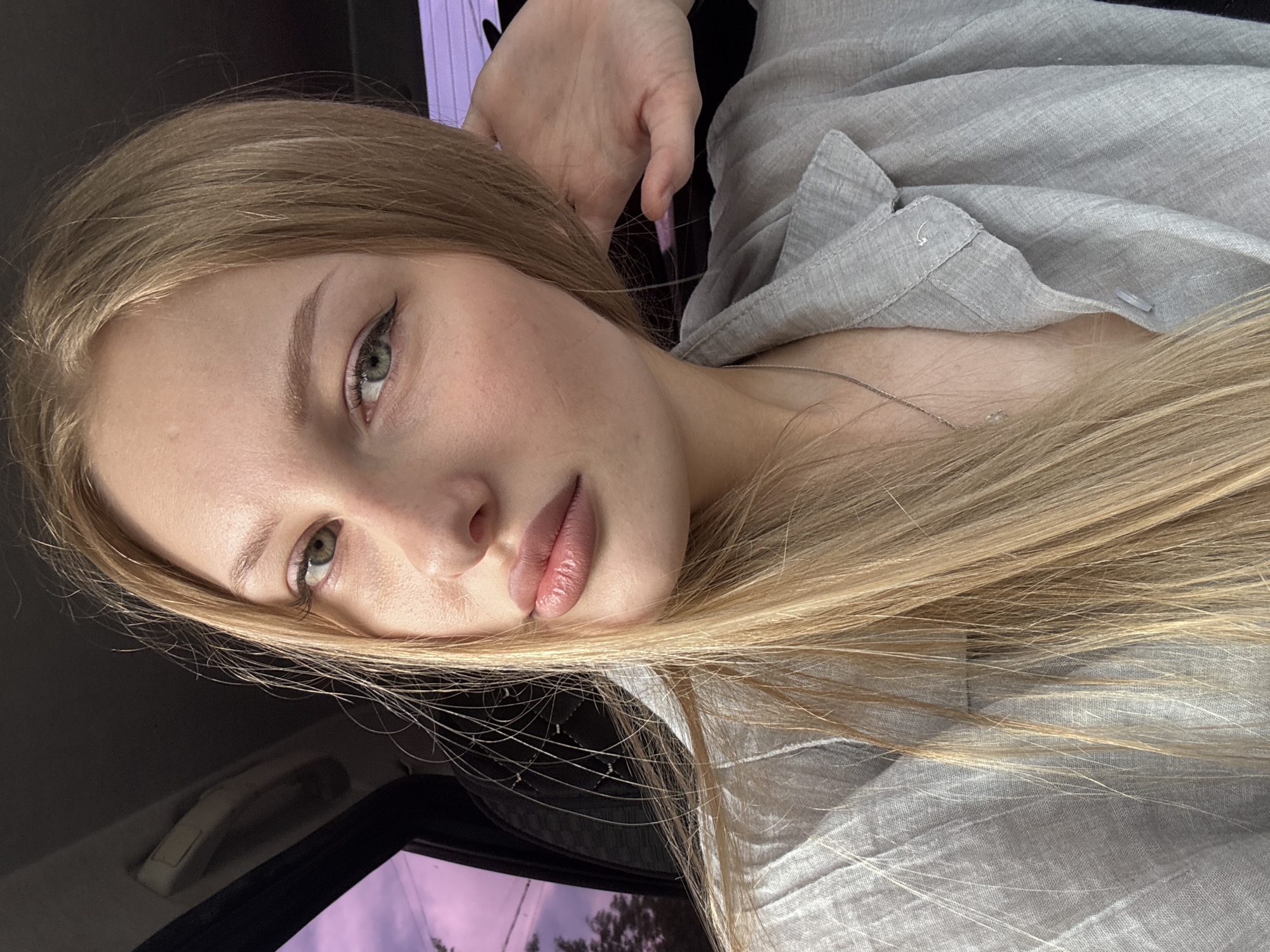 The image shows a close-up selfie of a young woman with blonde hair, fair skin, and light green eyes. She is wearing a light gray collared shirt. Her left hand is raised to the side of her head, and she is wearing a silver necklace. The background appears to be the interior of a car, with a window showing a blurred purple sky and trees. There is no identifiable information indicating the location is the city of Perm, Russia.