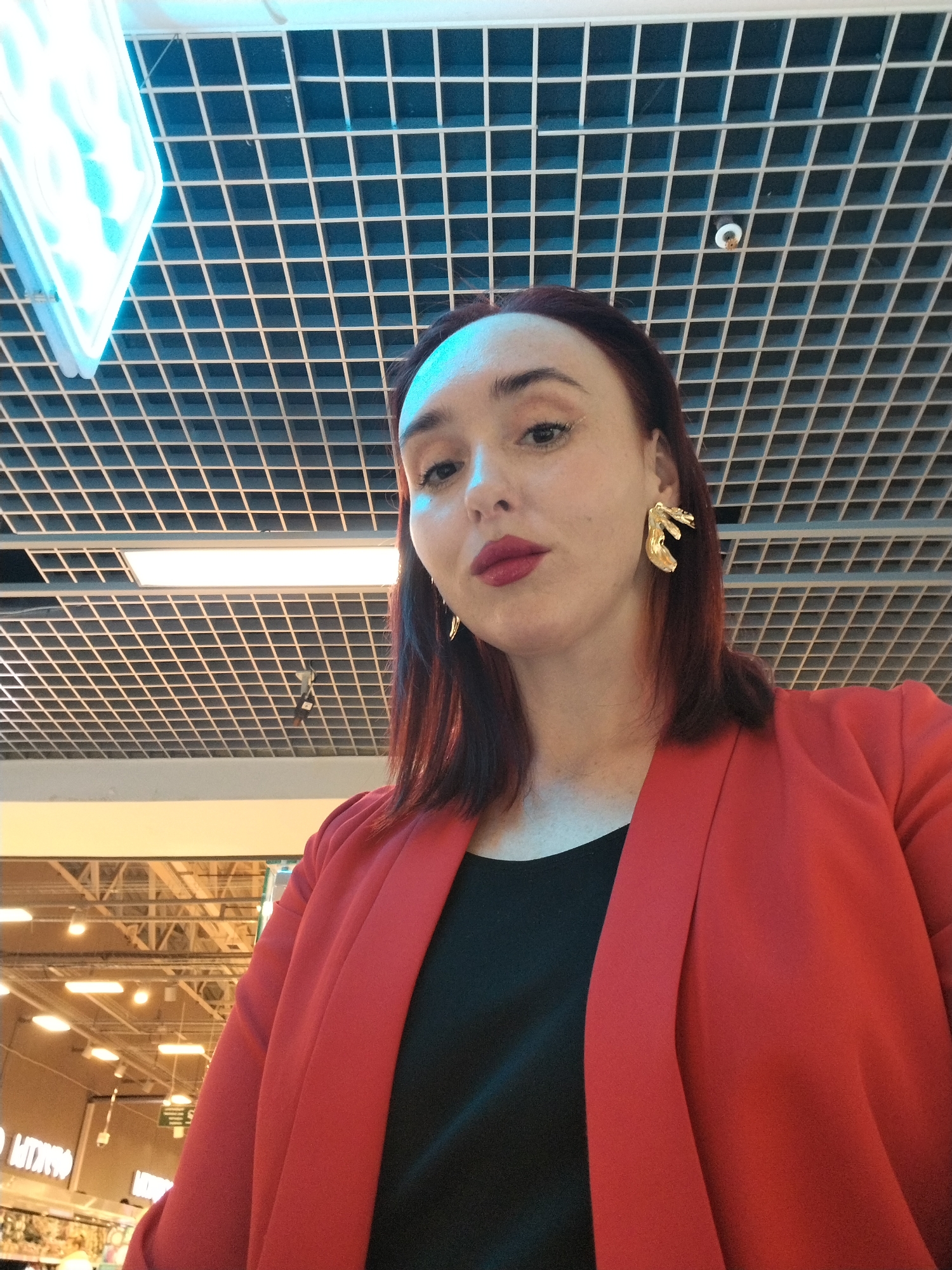 A woman stands in a shopping mall in Izhevsk, Russia. She wears a black shirt under a red blazer and large gold earrings. She has fair skin and dark red hair. The location appears to be a well-lit shopping mall, possibly a grocery store, due to the presence of overhead lighting and visible signage in the background. The woman is looking slightly upwards towards the camera. The ceiling is a grid-like structure, with lighting fixtures interspersed within.