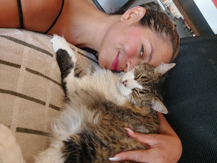 A woman with light skin and braided hair lies on a striped cushion with a fluffy, white and brown tabby cat. She has a lip piercing and is hugging the cat. The cat is lying on its back, with its eyes closed, appearing relaxed and content. A black mesh chair is visible behind the woman. The lighting is bright and natural.