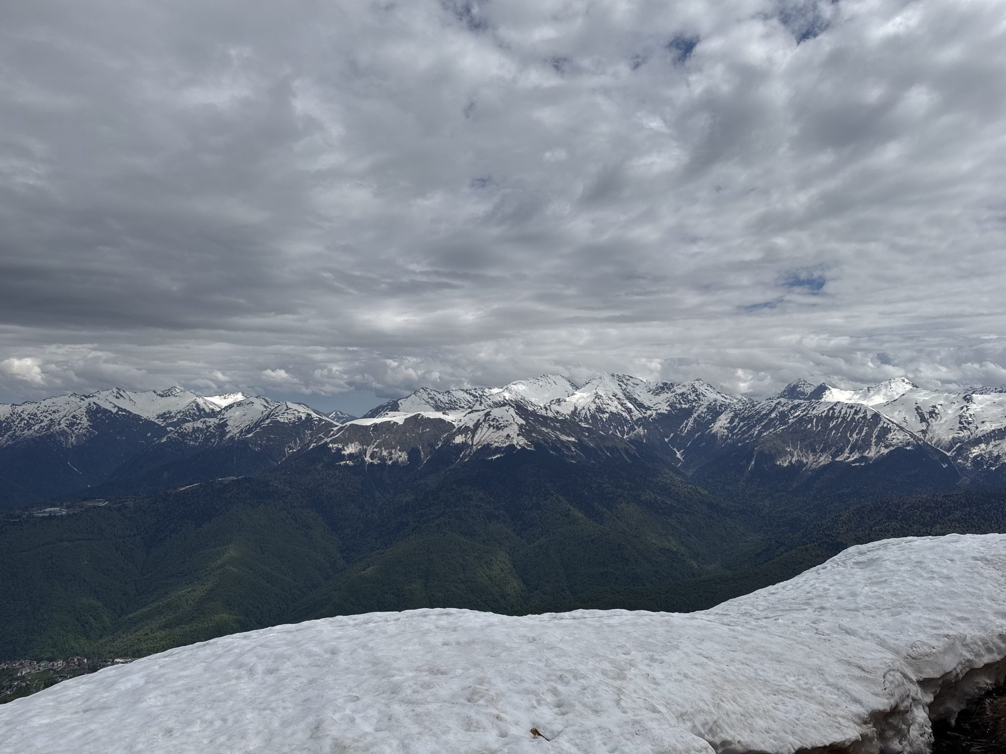 The image captures a mountain landscape in Estosadok, Russia. In the foreground, there's a snow-covered ledge. Behind it, a series of green, forested mountains rise, topped by snow-capped peaks. The sky above is filled with a heavy, grey cloud cover. A few buildings are visible at the bottom of the distant mountain range.