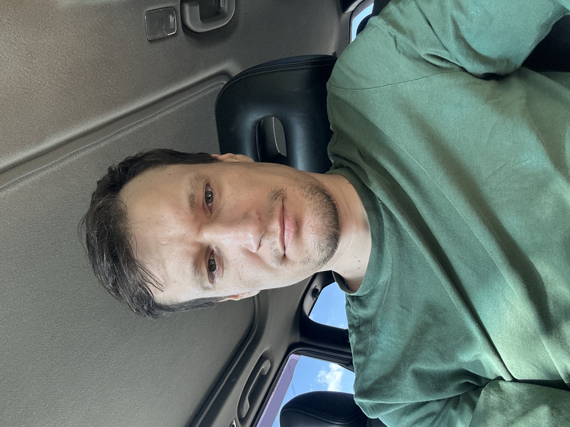 A man with short, dark hair and light skin is sitting in the driver's seat of a car, looking directly at the camera. He has a light beard and is wearing a green t-shirt. The car's interior is visible, including the headrest of the passenger seat and a sunroof. The sky and trees are visible through the car windows, suggesting an outdoor setting. There is no indication of the specific location within Makiv, Ukraine.