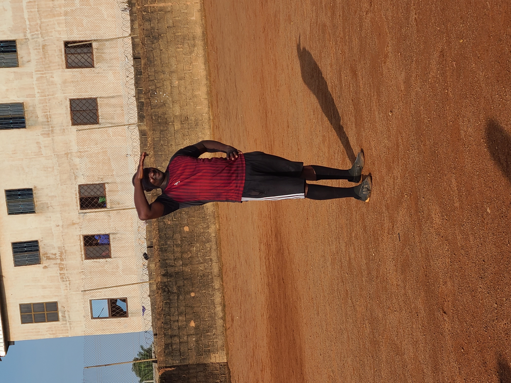 A man wearing a red and black striped soccer jersey and black shorts stands on a dirt field, shielding his eyes from the sun with his left hand. He is looking towards the right of the frame. In the background, there is a light-colored building with several windows, some of which are barred. A brick wall topped with barbed wire runs horizontally across the middle ground, separating the field from the building. The ground is covered in reddish-brown dirt and small stones. The image appears to have been taken outdoors, likely in Barakin Lerawi, Nigeria, given the architectural style and the warm, dry environment.