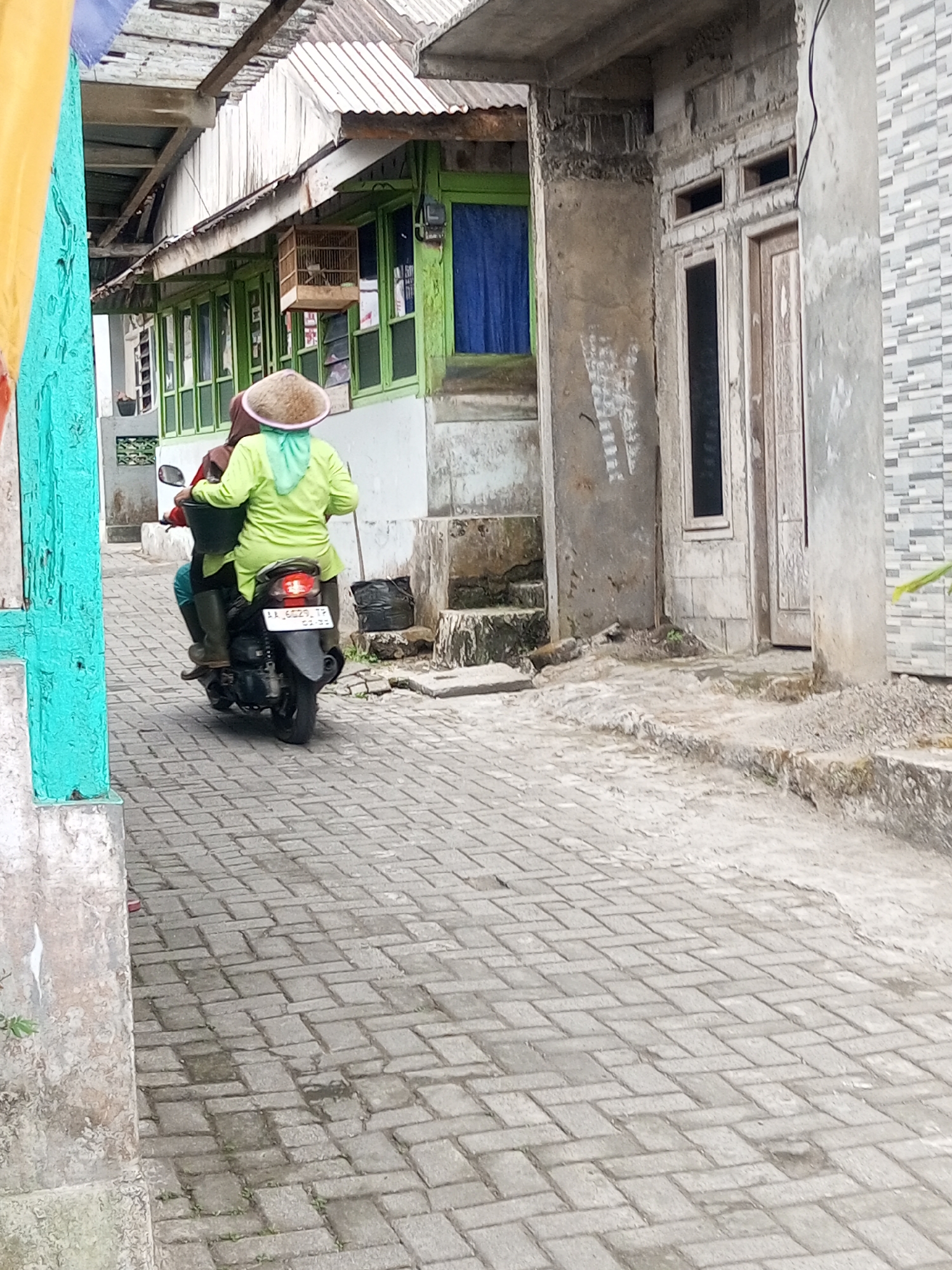 Two individuals are riding a motorcycle down a brick-paved alleyway in the city of Andongsili, Indonesia. The person in the back, wearing a light green long-sleeved shirt and a conical hat, is driving the motorcycle. The person in front, wearing a maroon shirt and a headscarf, is seated behind the driver. The motorcycle is black and appears to be moving forward. The alleyway is flanked by buildings on both sides, one with green-framed windows and a birdcage hanging outside, and the other with a textured concrete facade and wooden doors. A yellow and turquoise fabric hangs on the left side of the frame, partially obscuring the view of the buildings. The ground is paved with interlocking bricks, and there are some patches of dirt and debris on the sides. The lighting suggests it is daytime.