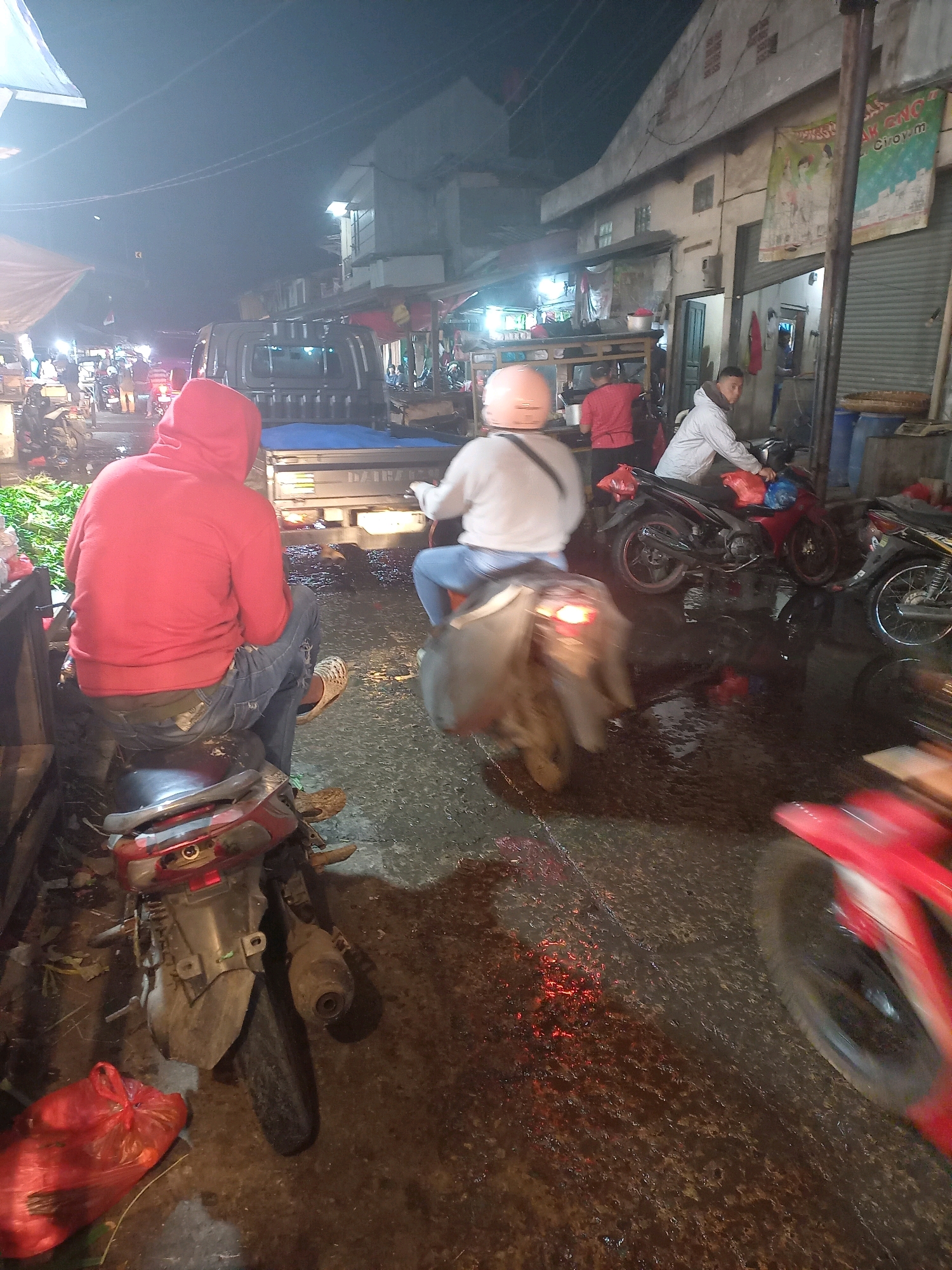 This is a nighttime, street-level, medium-shot photograph taken in what appears to be a busy market area in Bandung City, Indonesia. The scene is illuminated by artificial lights, creating a somewhat hazy and reflective atmosphere on the wet ground.
In the foreground, a person wearing a red hooded sweatshirt and blue jeans is seated on a motorcycle. They are facing away from the camera. To their left, a red plastic bag is on the ground.
Further into the scene, other individuals and motorcycles are present. One person is riding a motorcycle and is blurred due to motion. Another person in a light-colored jacket is also on a motorcycle, with a red plastic bag on the handlebars. A person in a red shirt is visible behind a market stall.
The background consists of buildings, some with signs that are partially visible. A large truck is parked in the middle ground. The overall environment suggests a commercial or street market setting, with various stalls and goods visible, though not clearly discernible. The wet pavement indicates recent rain or ongoing cleaning.