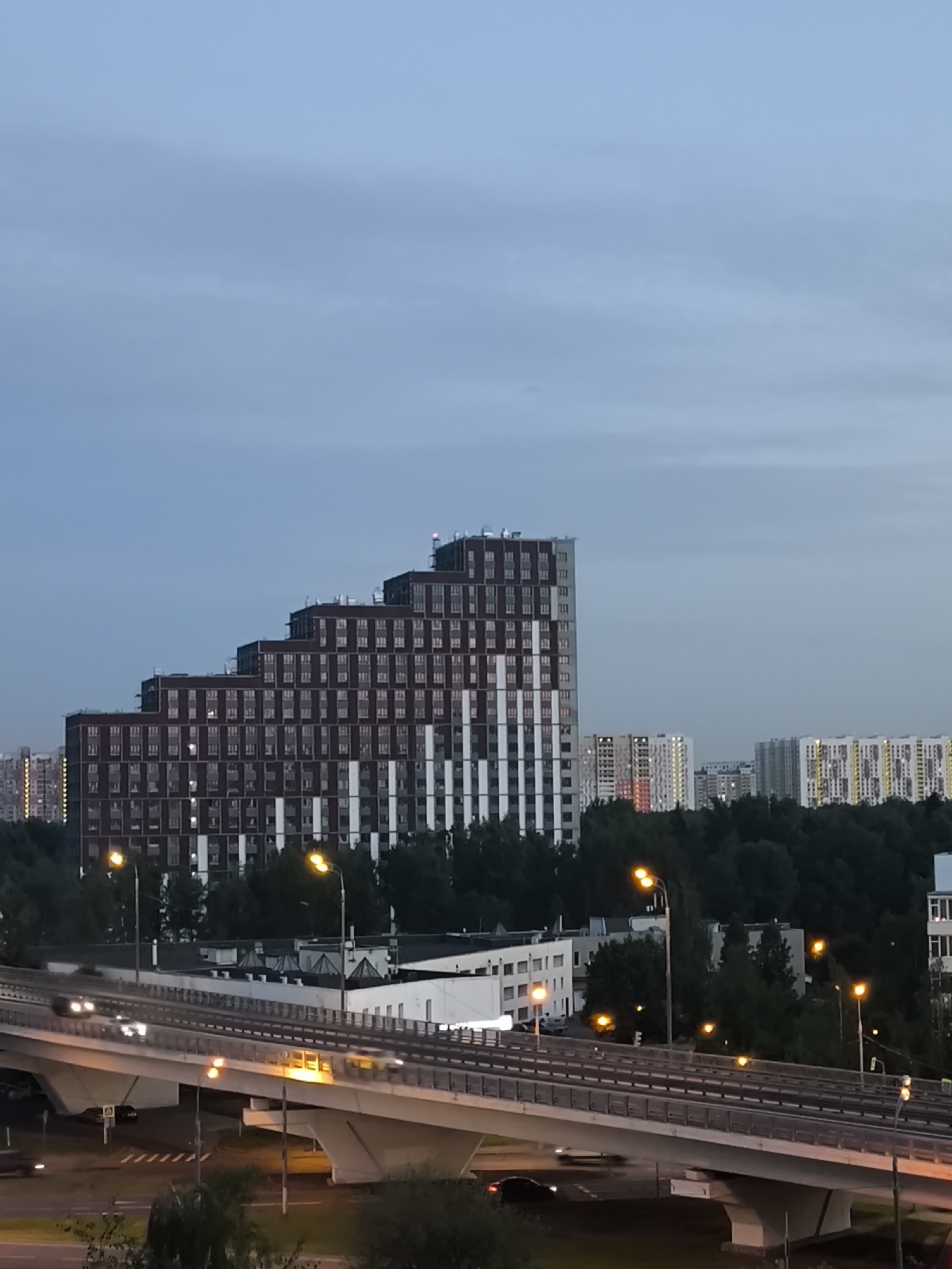 The image depicts an urban scene at dusk. A large, stepped residential building with dark red and white accents dominates the center-left of the frame. To the right of this building, several other multi-story buildings with illuminated windows are visible in the background, also surrounded by dark trees. In the foreground, an elevated multi-lane highway bridge spans across the scene, with several cars in motion, their headlights and taillights creating streaks of light due to motion blur. Streetlights with orange glows are present on the bridge and in the areas below. The sky is a muted blue-gray, indicating twilight. The overall impression is a view of a city during the transition from day to night.