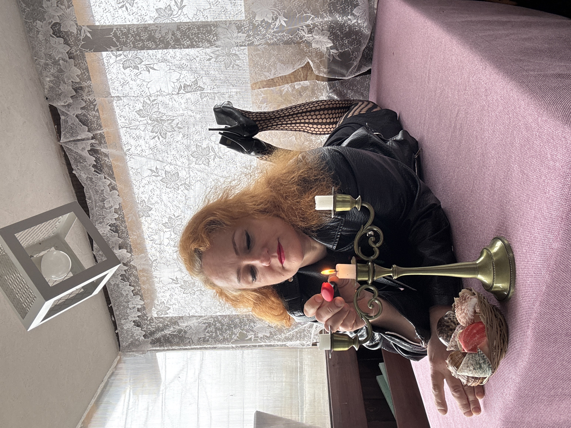 A woman with red hair and red lipstick is lying on a pink fabric-covered surface in a room in Chaikovsky, Russia. She is wearing a black leather jacket and fishnet stockings with black high heels. She is lighting a gold candelabra with white candles using a red lighter. Next to the candelabra is a small basket filled with seashells. A window covered with a lace curtain is in the background, and a modern white lantern-style light fixture hangs in the corner of the room.