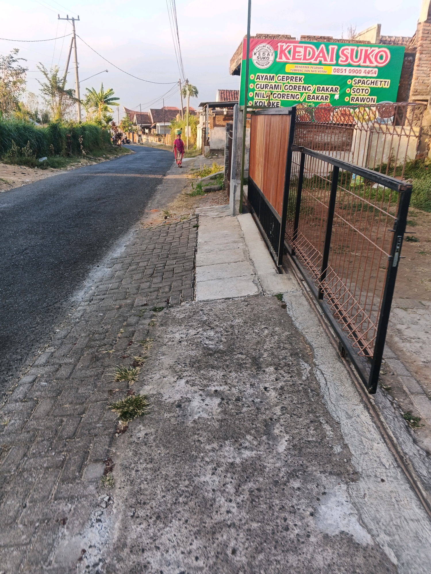 The image depicts a street scene in Bocek, Indonesia, with a focus on a roadside food stall named "Kedai Suko". The street is paved with asphalt, and to the left, there is a narrow brick walkway. In the foreground, to the right of the walkway, is a concrete area. Further down the street, a person in a red garment is walking away from the camera, carrying a bucket on their head. In the background, houses with terracotta roofs are visible, interspersed with lush green vegetation. The Kedai Suko stall itself is characterized by a large green banner advertising various food items like "AYAM CEPGEK", "CURAMI GORENG/BAKAR", "NILAI GORENG/BAKAR", "KOLOKE", "SPAGHETTI", "SOTO", and "RAWON". A sliding metal gate with wooden panels is positioned in front of the stall. The overall atmosphere suggests a rural or suburban setting, possibly in the morning or late afternoon given the soft lighting.