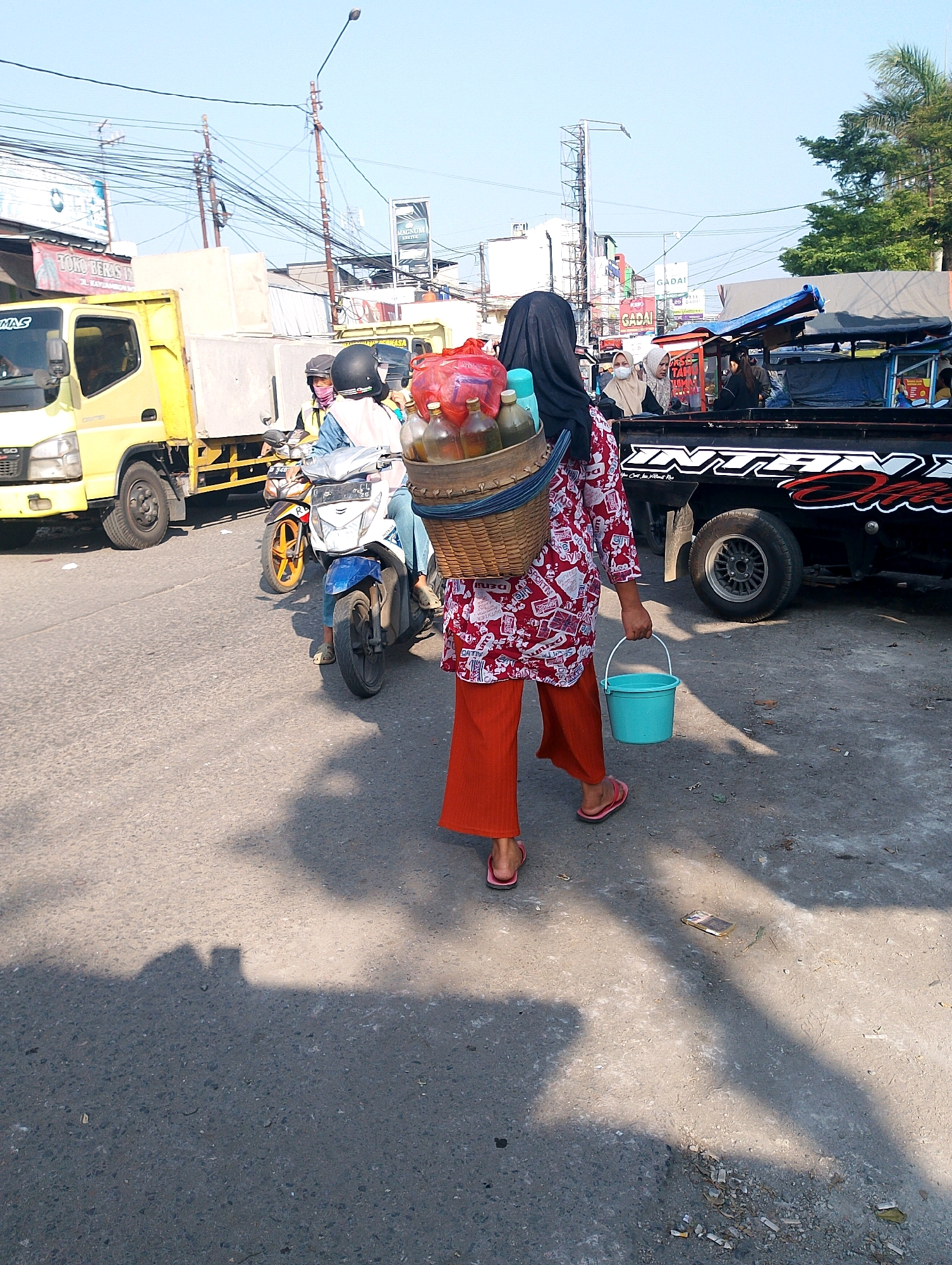 A medium shot shows a person from the back wearing an orange headscarf, a red and white patterned shirt, and orange pants. They are carrying a woven basket filled with bottles and a blue bucket in their right hand. The person is walking on a paved road with a shadow falling across it. In the background, there are several vehicles, including a yellow truck and a black truck with a logo. There are also overhead wires and streetlights. The scene appears to be a busy street in Lembang, Indonesia, possibly near a market or roadside stall, indicated by the presence of vendors and parked vehicles. The sky is clear and blue.