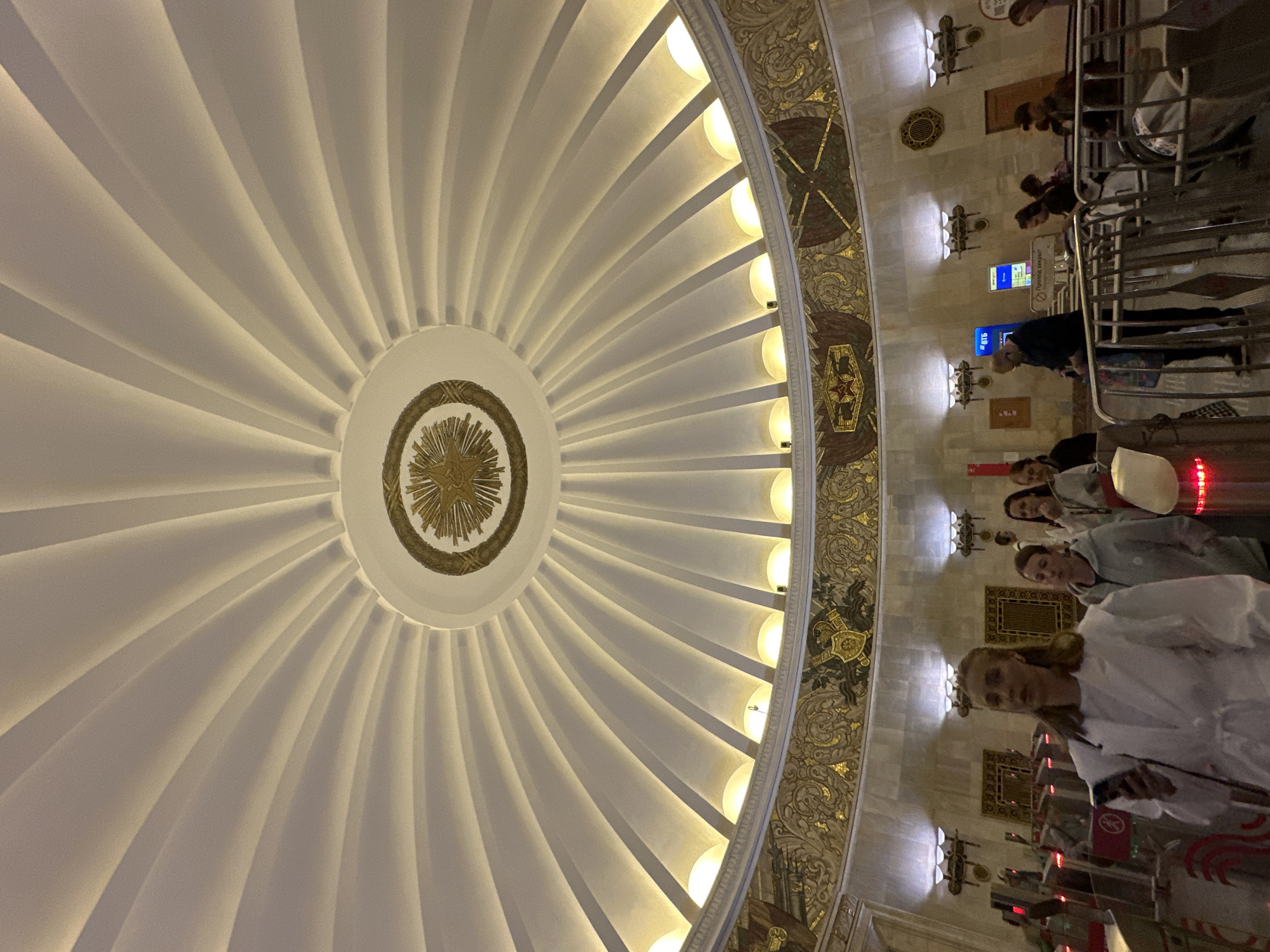 This is a photograph taken inside a metro station in Moscow, Russia, likely at a station with a domed ceiling and ornate decoration. The focus is on the impressive, sunburst-like white dome of the ceiling, illuminated by integrated lighting that creates dramatic shadows. Below the dome, a decorative frieze runs along the upper part of the walls, featuring golden patterns and Soviet-era symbols such as stars and banners.
In the lower part of the image, several people are seen interacting with a turnstile system, suggesting they are entering or exiting the metro platforms. One woman in the foreground, wearing a white robe-like garment and holding a smartphone, is looking towards the camera. Other individuals, dressed in casual attire, are also visible, some waiting in line, others walking. The overall environment appears to be a grand, well-lit public space characteristic of some of Moscow's historic metro stations.