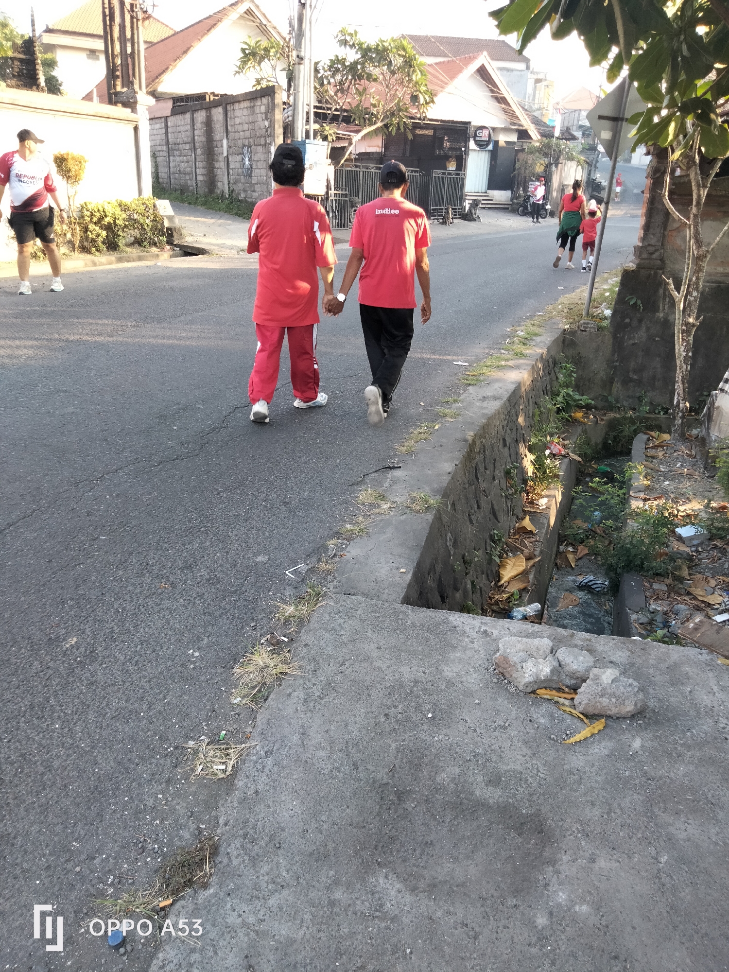 In Denpasar, Indonesia, on a paved street adjacent to a concrete drain filled with debris, two individuals walk side-by-side. Both are wearing red tops and dark pants, with one carrying a red bag. The person on the left is wearing a black cap and a red and white shirt with "REPUBLIK INDONESIA" printed on it, along with black shorts and white sneakers. Further down the street, another person in red is seen walking away from the camera. The environment shows residential buildings and vegetation.