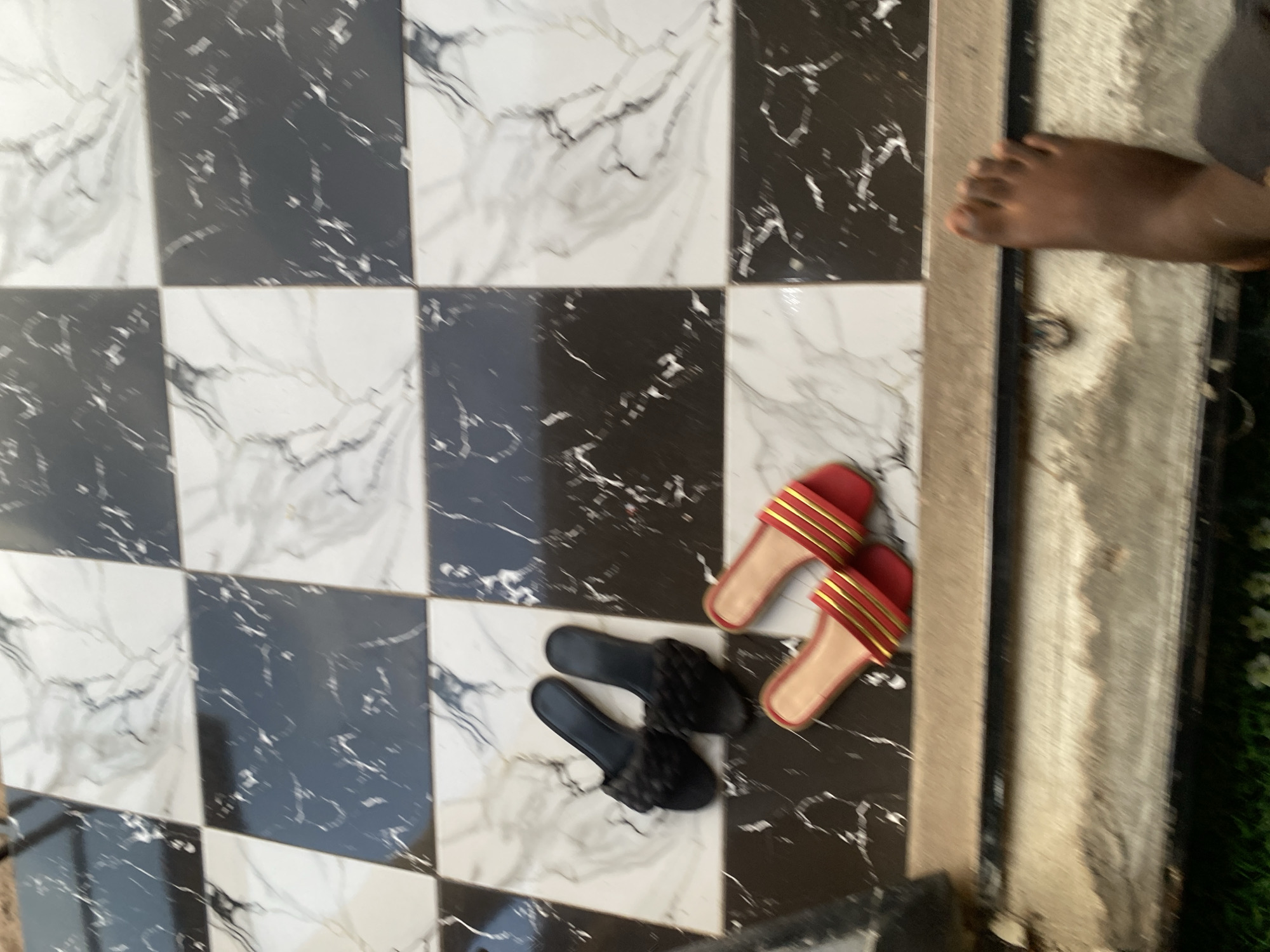 An outdoor, eye-level shot captures a checkerboard tiled floor in Jauro Yinu, Nigeria. The tiles are a mix of black and white with marble-like veining. Two pairs of women's sandals are present. One pair is black with a woven texture, and the other is red with gold stripes. In the foreground, a bare foot, belonging to an adult, is visible from the ankle down. The foot is dark-skinned. The surrounding area includes what appears to be a threshold with some debris.