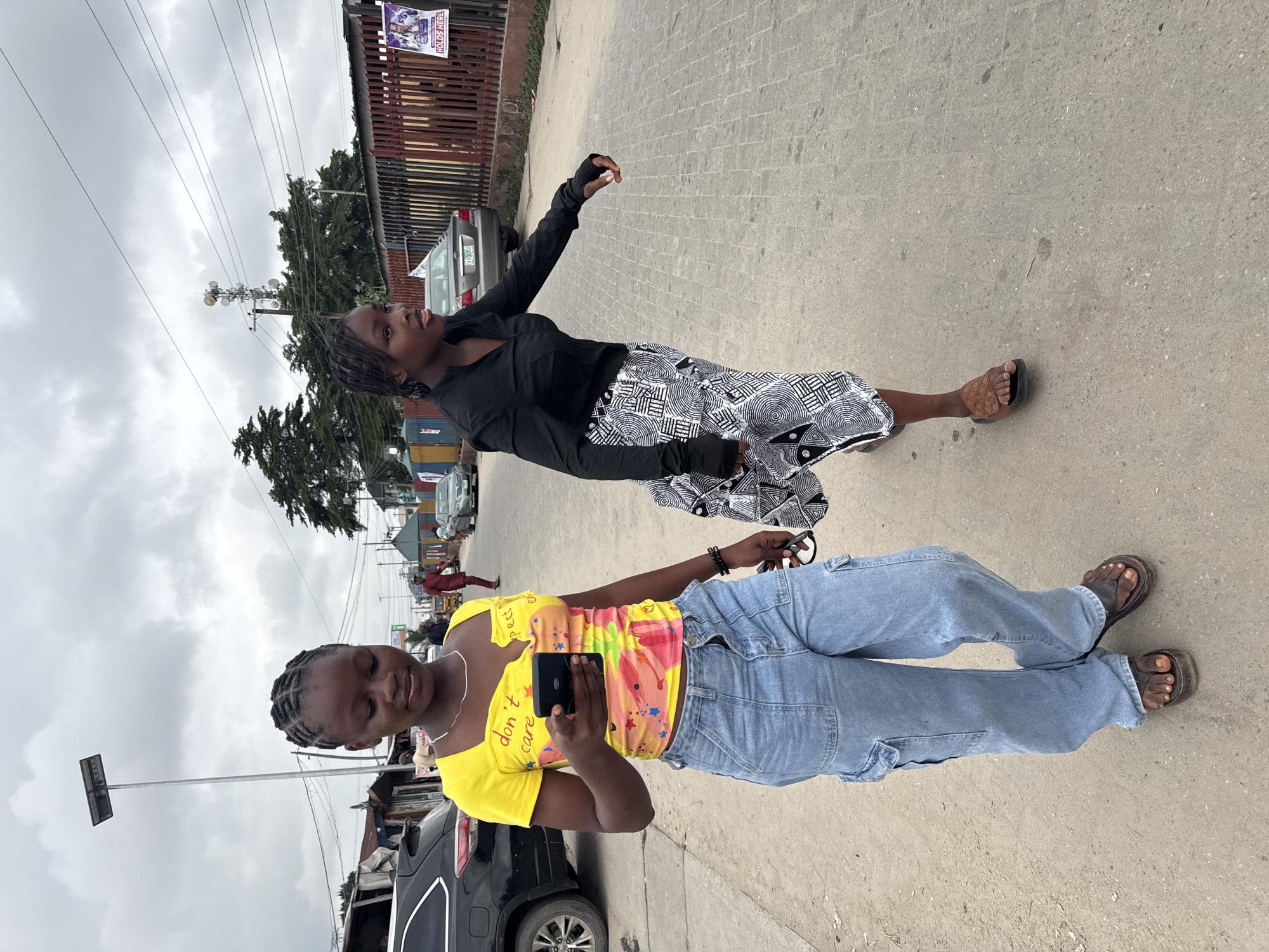 Two young Black women are walking on a paved street in Ado, Nigeria. The woman on the left, in the foreground, wears a yellow, scoop-neck t-shirt with a colorful floral and star pattern and the words "don't care" printed on it. She has her hair braided into cornrows and is looking down at her phone, which she holds in her right hand. She is wearing light blue cargo pants and dark brown sandals. She is holding a pair of dark-rimmed glasses in her left hand.
The woman on the right, slightly behind and to the left of the first woman, wears a black long-sleeved shirt and a black and white patterned skirt. Her hair is also braided into cornrows. She is looking to her right, with her mouth slightly open. She is wearing brown slide sandals.
The environment is an urban street scene. In the background, there are buildings, fences, parked cars, and a utility pole with a solar panel at the top. There are also power lines and some trees visible. The sky is overcast. A poster is visible on a fence to the right. The pavement is made of interlocking grey bricks and has some dark stains.