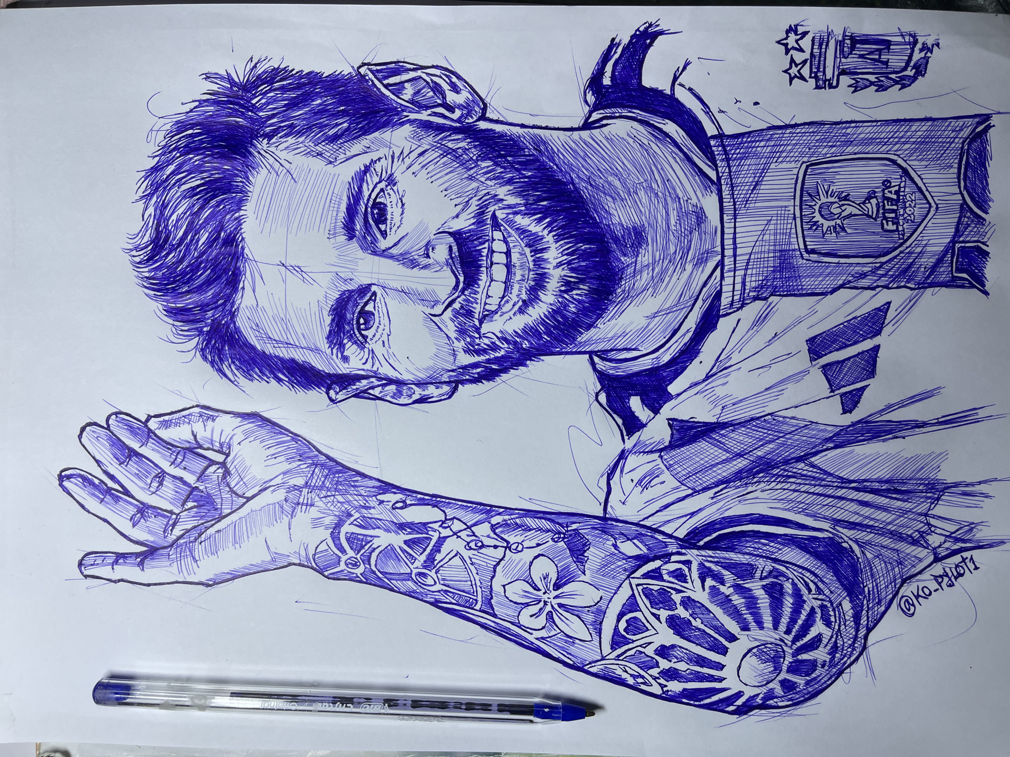 A pen and ink drawing on a white surface depicts a portrait of Lionel Messi. He is shown from the chest up, with his left arm raised and hand open, palm facing forward. Messi has a beard and short, tousled hair. He is wearing a jersey with a FIFA World Cup 2022 badge and what appears to be an Adidas logo on the chest. There are also decorative tattoos visible on his left arm. A blue ballpoint pen is placed vertically on the left side of the drawing. The background is plain white, with some stray ink marks and sketching lines visible. The drawing is signed "@KO_pblot1" in the lower left corner. There is no information about the location in the city of Jos, Nigeria, within the image or its context.