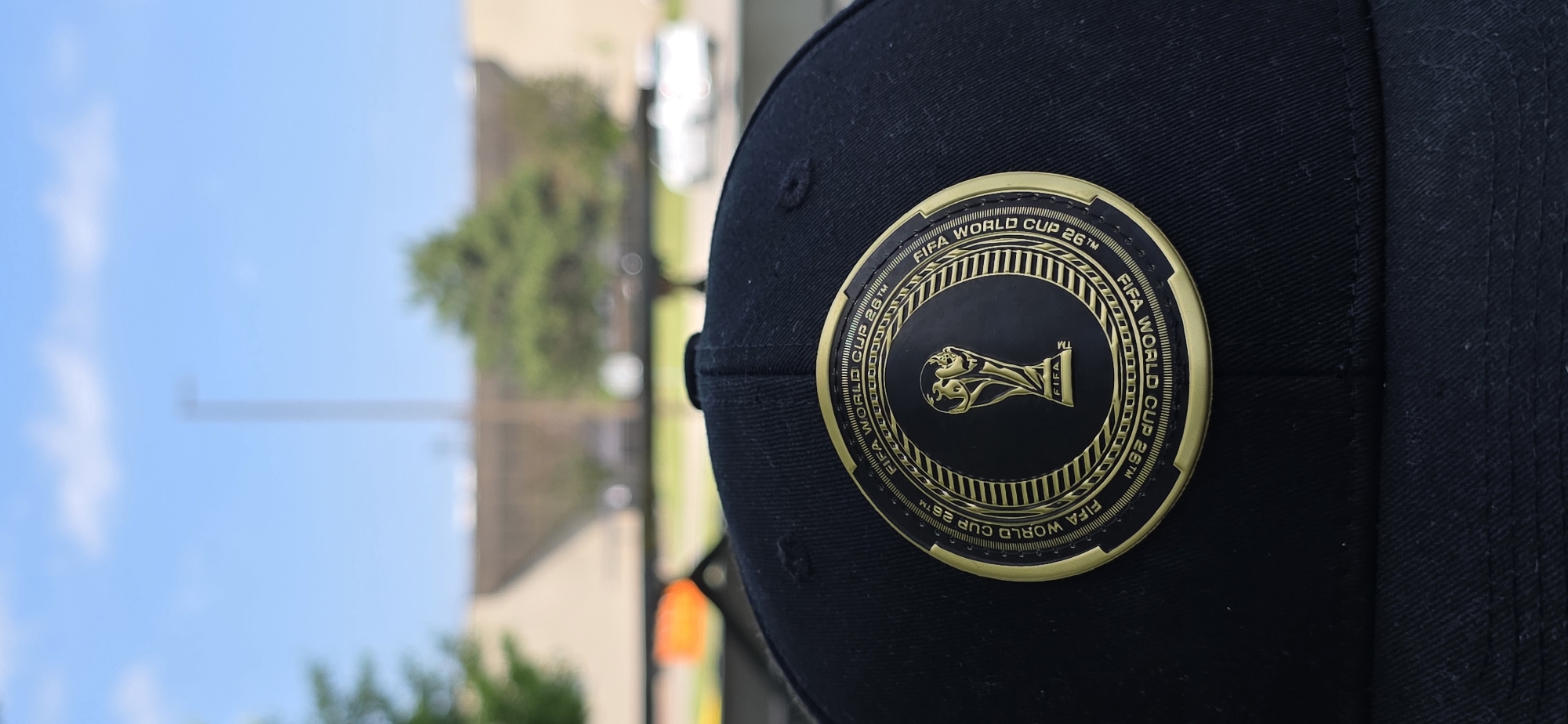 A close-up, eye-level, vertical outdoor shot captures the top of a black baseball cap. The cap features a circular, gold and black emblem of the FIFA World Cup trophy, surrounded by text reading "FIFA WORLD CUP 26TM". The cap is situated in the foreground, with a blurred background of a bright blue sky with white clouds, and a distant, blurred cityscape of buildings and trees. The overall impression is of an outdoor setting, possibly a park or public area, with a focus on the cap's emblem. The exact location in Pickering, Canada is not discernible from the provided image.