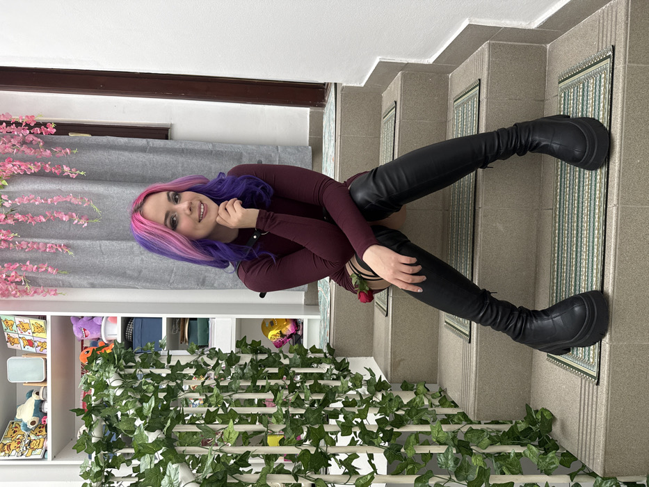 An indoor scene depicts a young female with distinct pink and purple hair, seated on the third step of a light-colored staircase. She is wearing a dark red, long-sleeved dress, black over-the-knee platform boots with thick soles, and a black choker-style collar with a metal ring. A single red rose is tucked into a black garter visible on her right thigh. Her right hand is near her chin, and she is smiling directly towards the viewer. Each step of the staircase is fitted with a small, rectangular mat featuring a green and white striped pattern. To the left of the staircase, a white shelving unit containing various items, including plush toys and framed pictures, is partially visible behind a white railing adorned with artificial green ivy vines. To the right, a grey curtain covers a window or opening, with strands of pink artificial wisteria-like flowers suspended above it. The setting appears to be an interior space, likely residential or private. No external landmarks or distinguishing features are present to pinpoint a specific location within the City of Zagreb, Croatia.