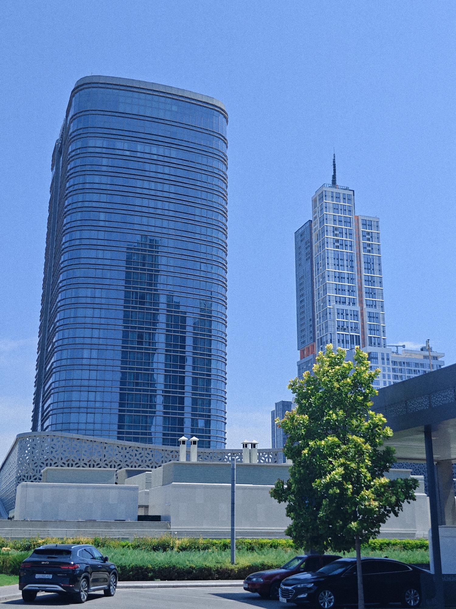 The image captures a sunny day in Tashkent, Uzbekistan, with a focus on modern architecture. A tall, curved skyscraper with a blue glass facade dominates the left side of the frame. To its right, a slender, multi-story skyscraper with a spired top stands against a clear blue sky. In the foreground, a dark SUV is parked on a paved surface. Behind the SUV, a lush green lawn borders a row of shrubs and decorative patterned walls. A vibrant green tree stands in the middle ground, partially obscuring other buildings. The scene suggests a well-maintained urban environment with contemporary design elements.