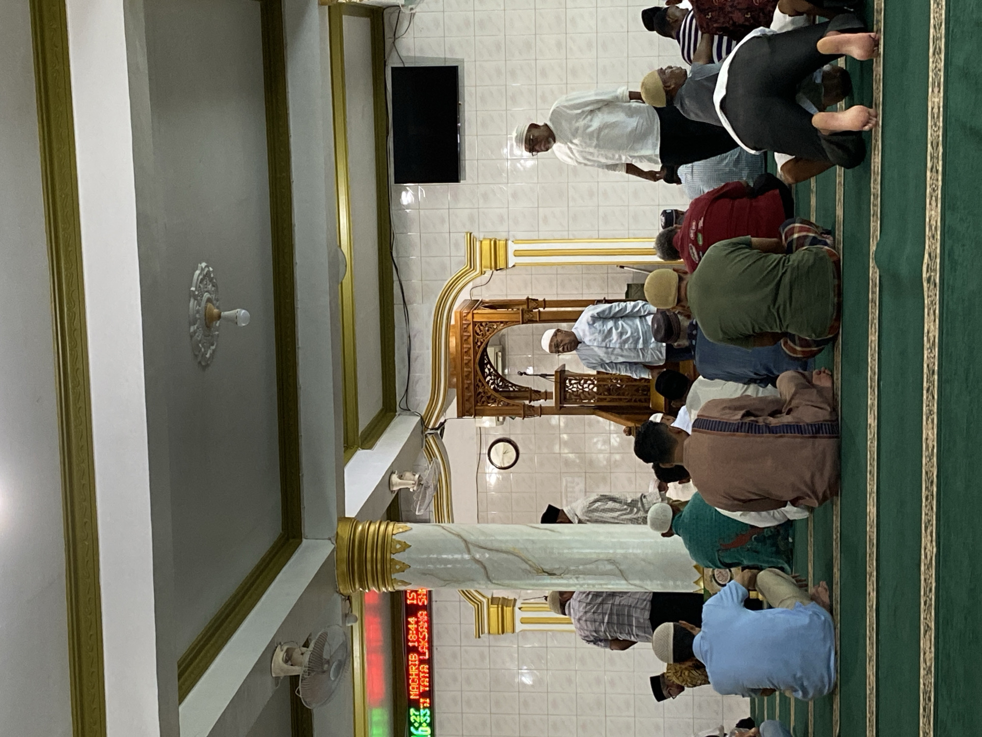 This image captures a group of Muslim men engaged in congregational prayer (salat) inside a mosque in Lhokseumawe, Indonesia. The men are arranged in rows on green prayer mats adorned with gold stripes, performing either prostration (sujood) or a sitting posture (jalsa/tashahhud), indicating different stages of the prayer ritual. They wear a variety of attire, including traditional shirts and trousers, some with skullcaps (kufis or songkoks).
The setting is an indoor prayer hall characterized by white tiled walls and a white ceiling with elaborate gold trim and decorative elements. A large, ornate wooden mihrab or minbar structure, typically used by the imam, is visible at the front, indicating the direction of prayer (Qibla). A prominent white marble column with gold accents stands in the foreground.
Several objects are present: a black, rectangular wall-mounted television screen, a traditional round analog clock on the wall, and multiple small wall-mounted fans, suggesting a warm climate. A decorative ceiling fixture, possibly for lighting, is also visible.
A digital display panel in the lower left corner shows visible text. The red segment displays "NAGHRIB 18:44 IS" and "06:27", while a green segment shows "TATA LAKSAMA SH". "NAGHRIB" refers to the Maghrib (sunset) prayer, and "18:44" is likely its time, with "IS" potentially indicating Isya prayer. The "06:27" could be the current time or Fajr prayer time, suggesting the image was taken in the evening, possibly during or shortly after Maghrib prayer. The overall atmosphere appears solemn and devotional.